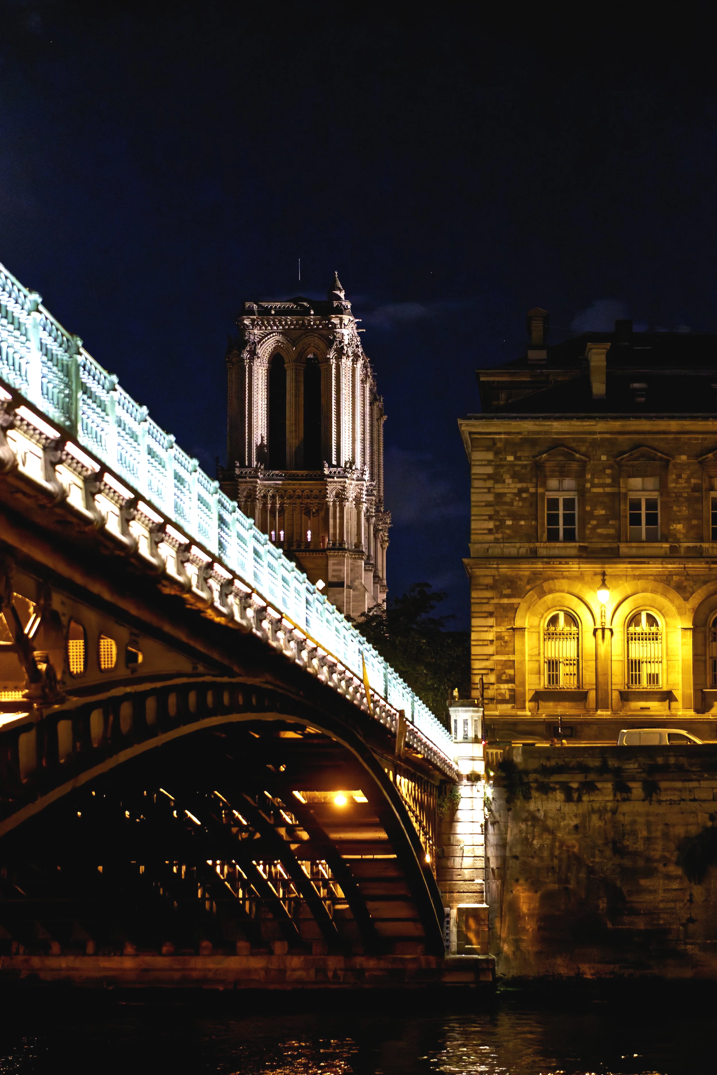Nighttime view of a bridge illuminated with lights, with a historic building and the Notre-Dame Cathedral in Paris, France in the background.