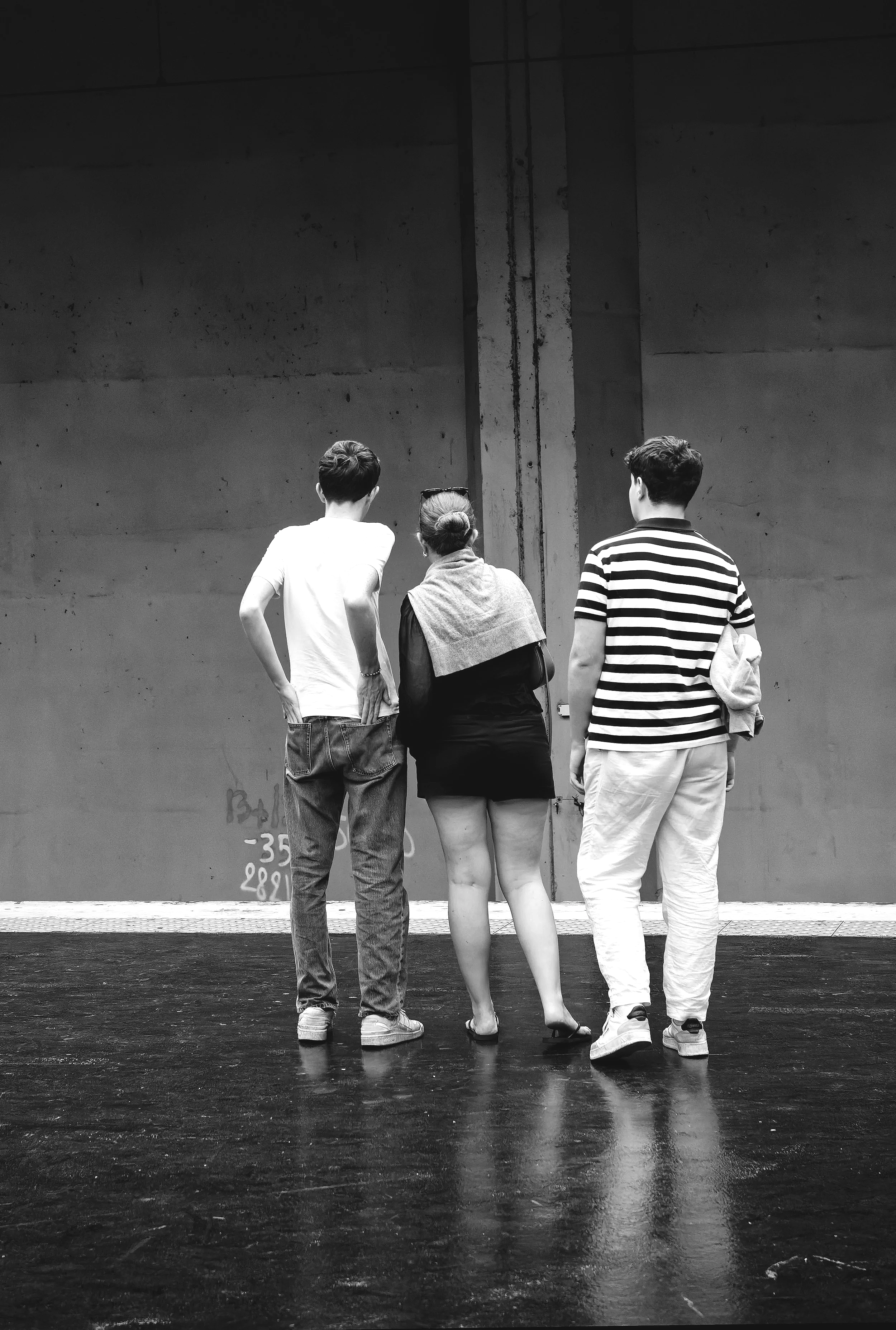 Three people standing together in an indoor setting, viewed from the back. One person wears a striped shirt and light pants, another in a white T-shirt and jeans, and the third in a black outfit with a scarf. The background is a plain concrete wall.