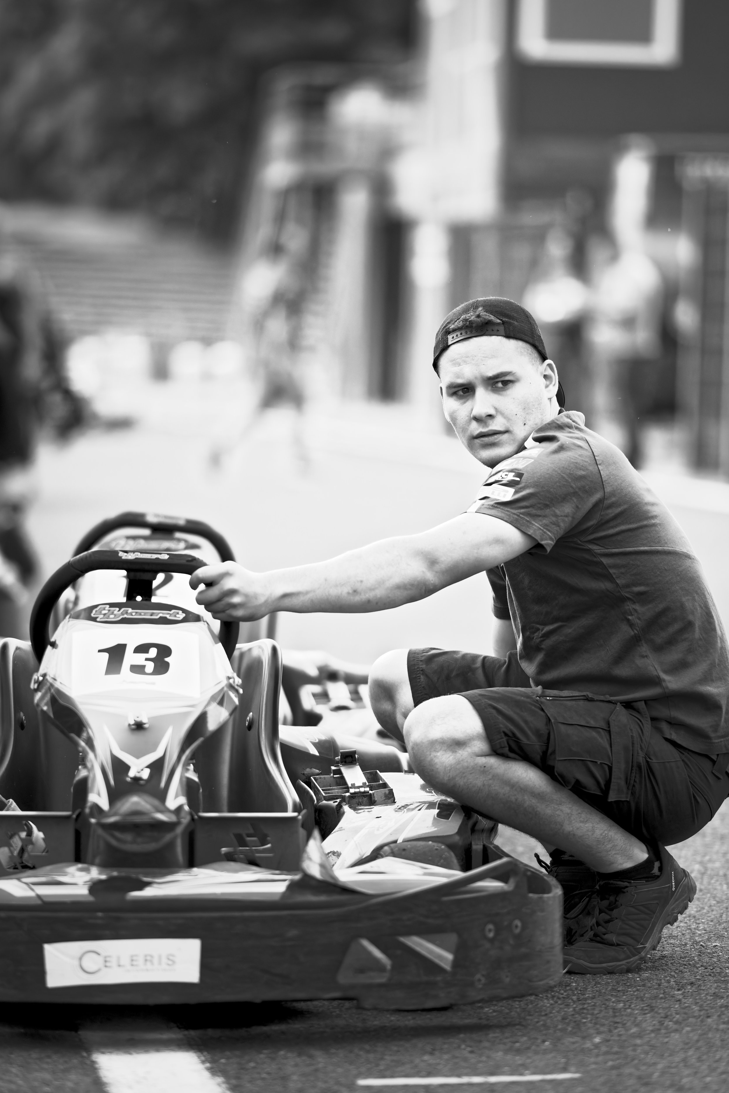 A young man with a cap squatting next to a go-kart with the number 13, on a street with blurred background.