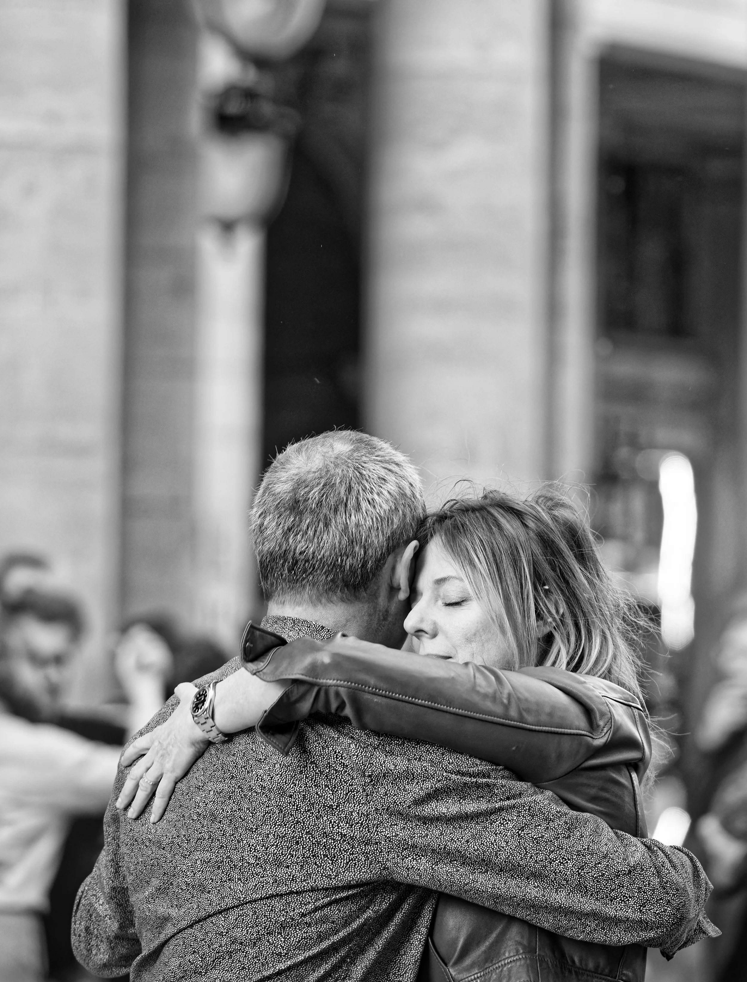 A black and white photo of a man and woman embracing, with the woman’s eyes closed and a content expression, in a social or public setting.