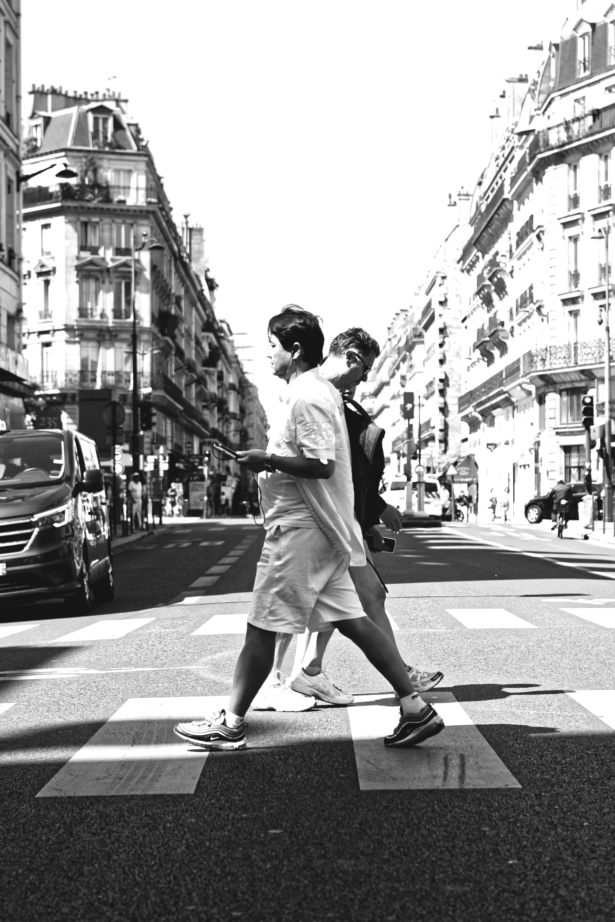 Black and white photo of two people walking across a city street with buildings on either side, cars parked along the curb, and pedestrians in the background.