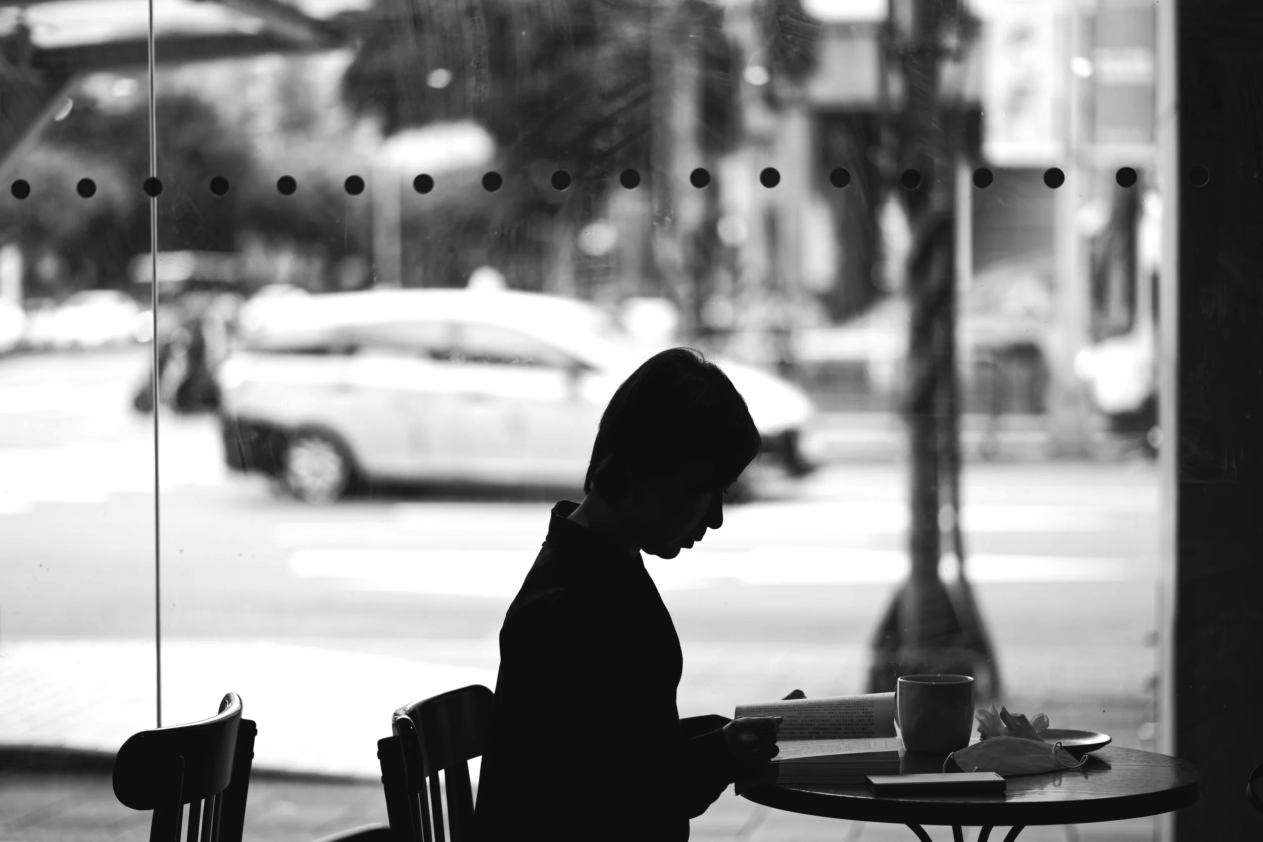 A woman sitting alone in a cafe, reading a book. The scene is viewed through a glass window with cars and trees outside, in black and white.