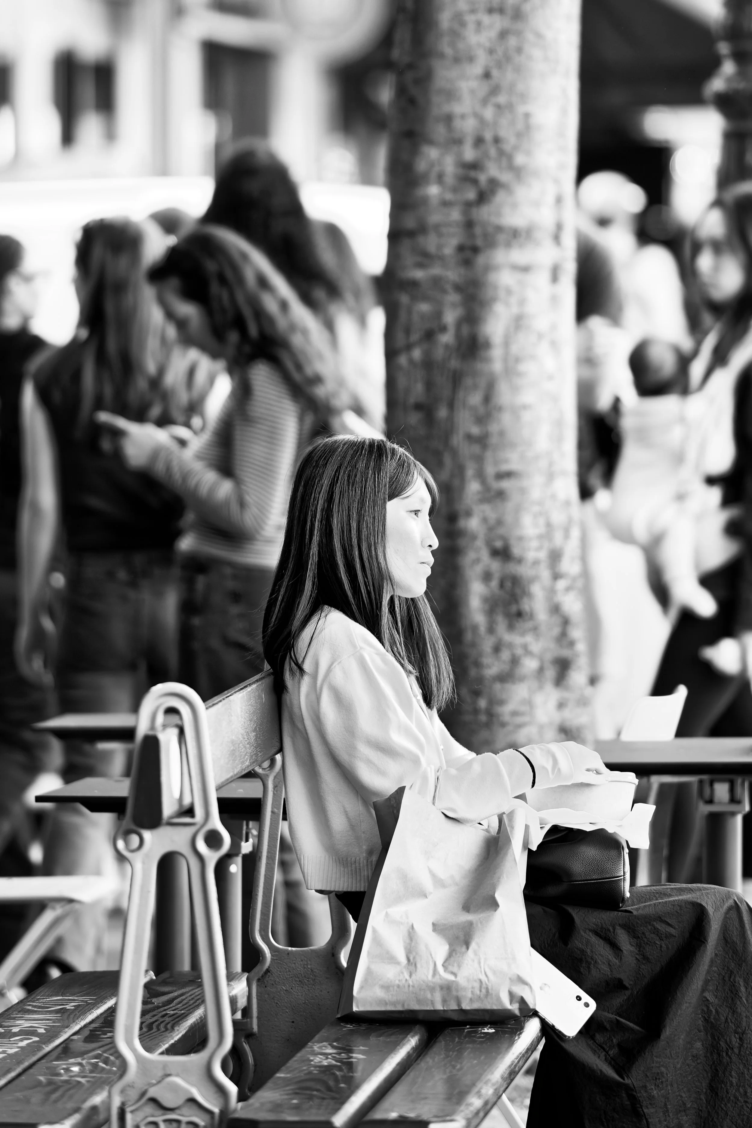 A woman sitting on a bench on a city street, surrounded by a crowd of people, with a tree in the background.