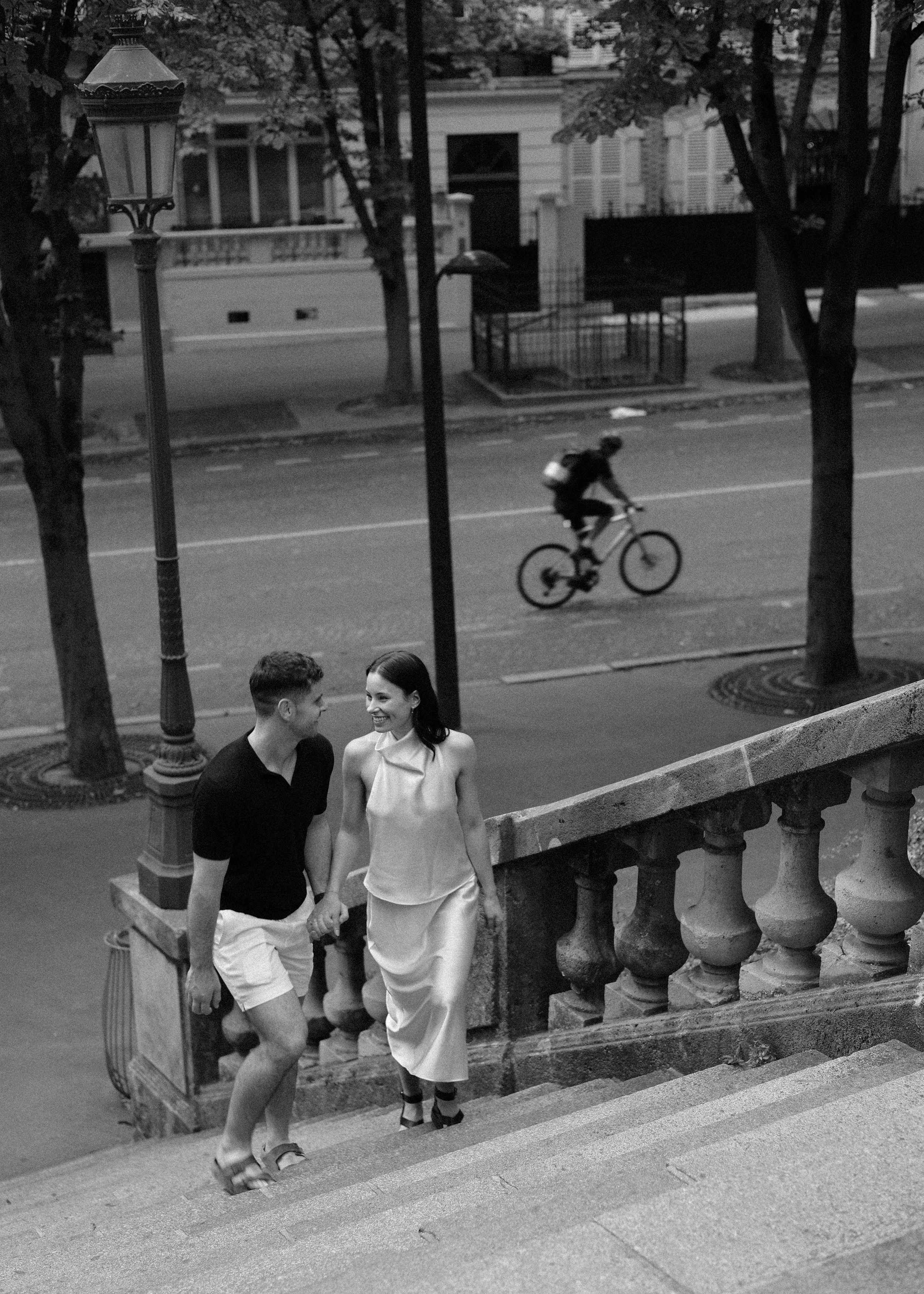 A black-and-white photo of a young couple walking up a stone staircase on an urban street at night, holding hands and smiling at each other. A cyclist is riding on the street behind them.