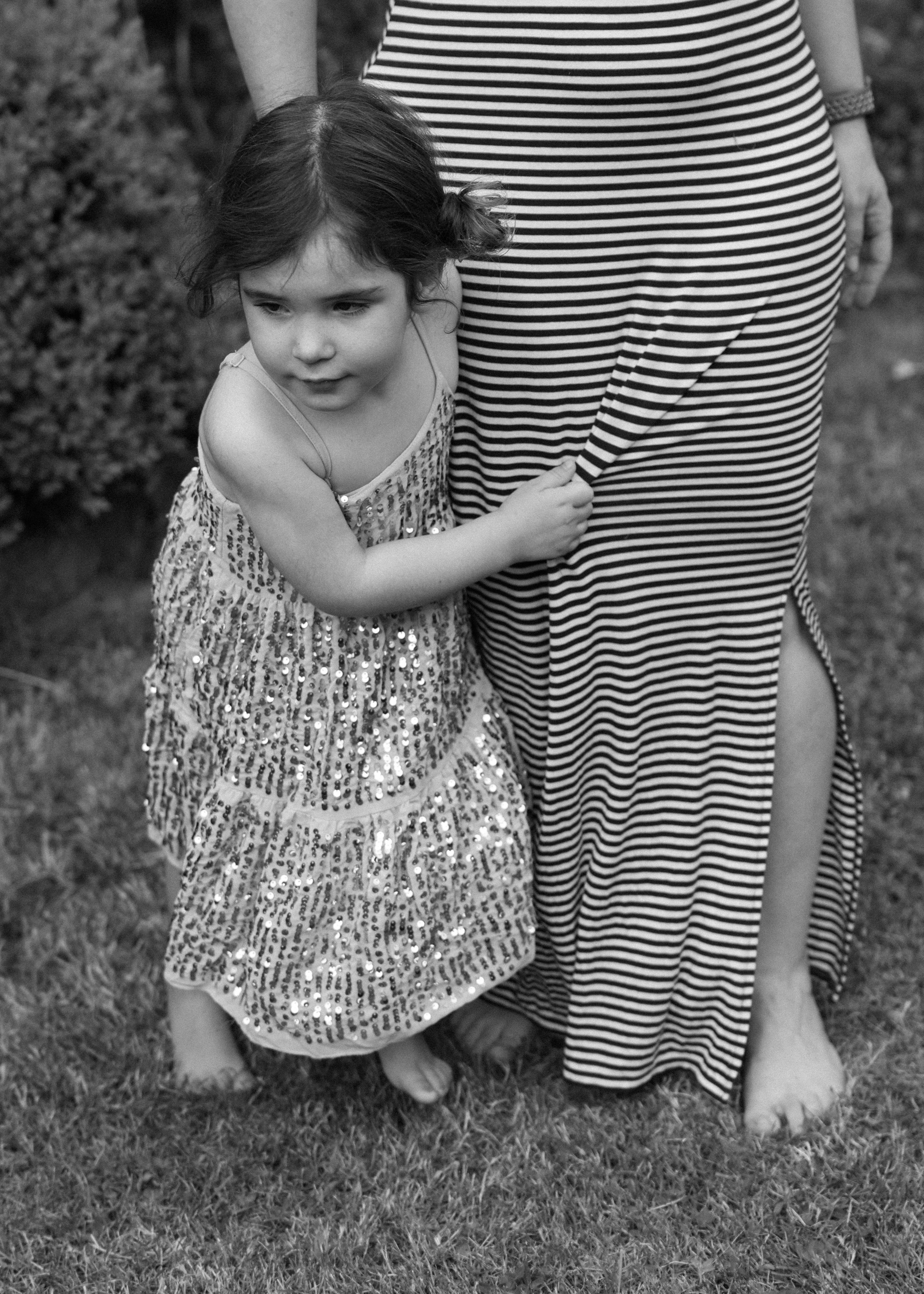 A young girl with dark hair wearing a sequined dress, hugging an adult in a striped dress with a slit, standing on grass in a garden.