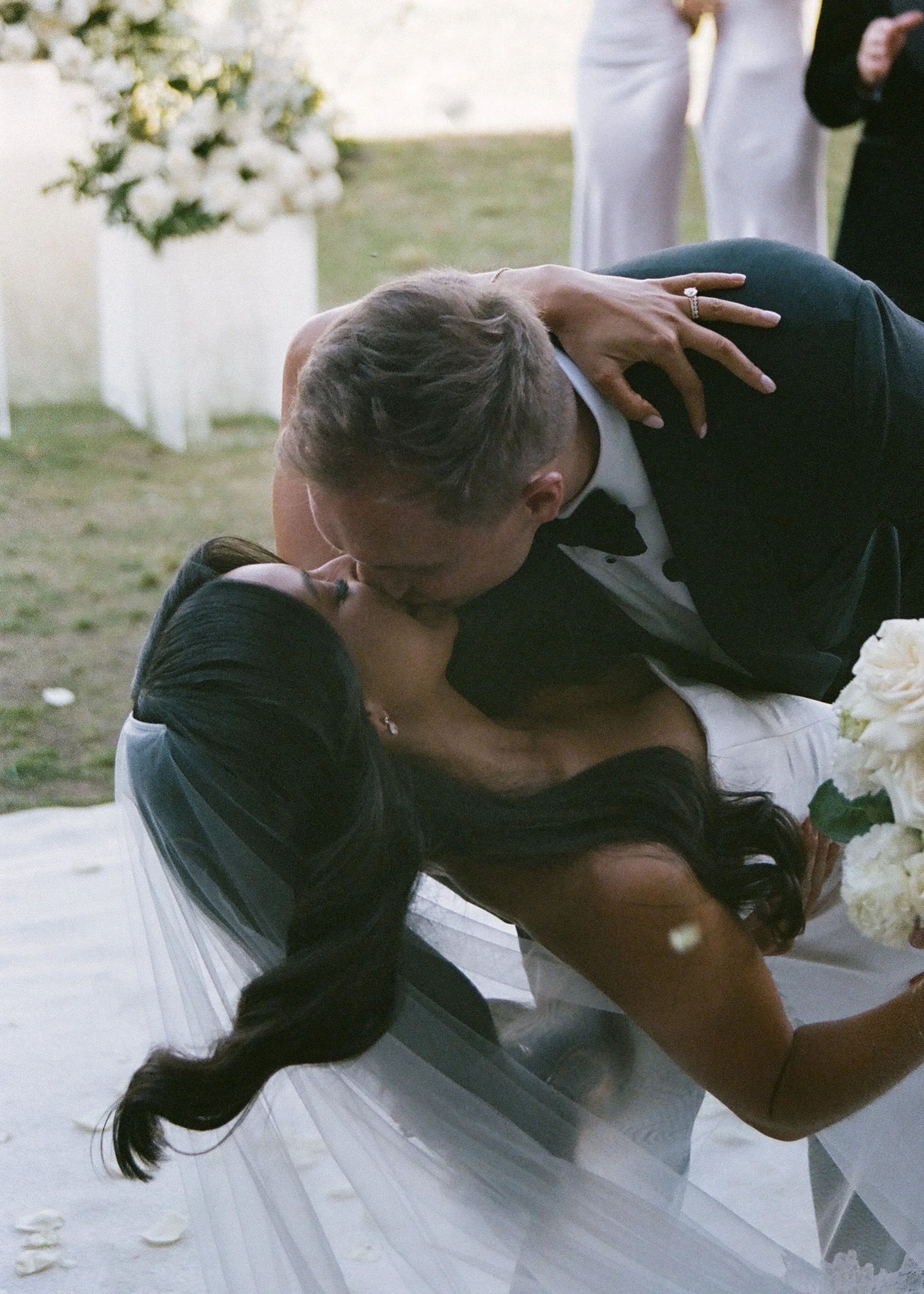 A bride and groom share a kiss at their wedding reception, with the bride wearing a veil and holding a bouquet of white flowers, and an outdoor setting with guests in the background.