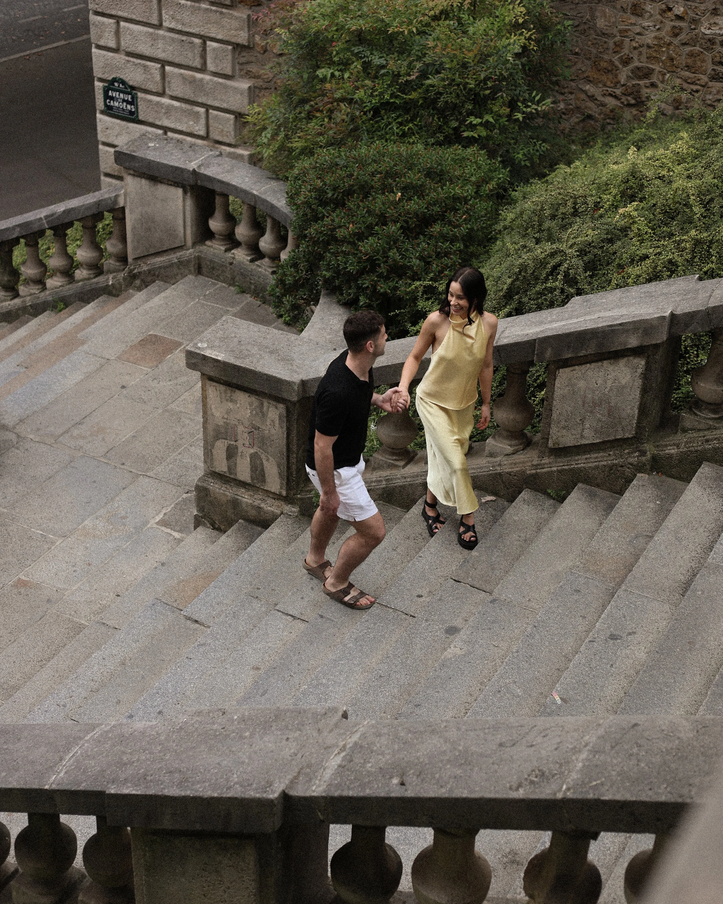 A man and woman holding hands while walking up stairs outside, surrounded by greenery and stone railings.