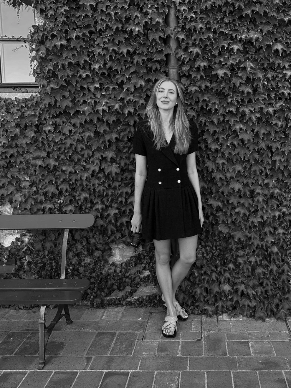 A woman with long hair, wearing a black dress, stands in front of a wall of ivy, holding a camera in her right hand, with a bench to her left.