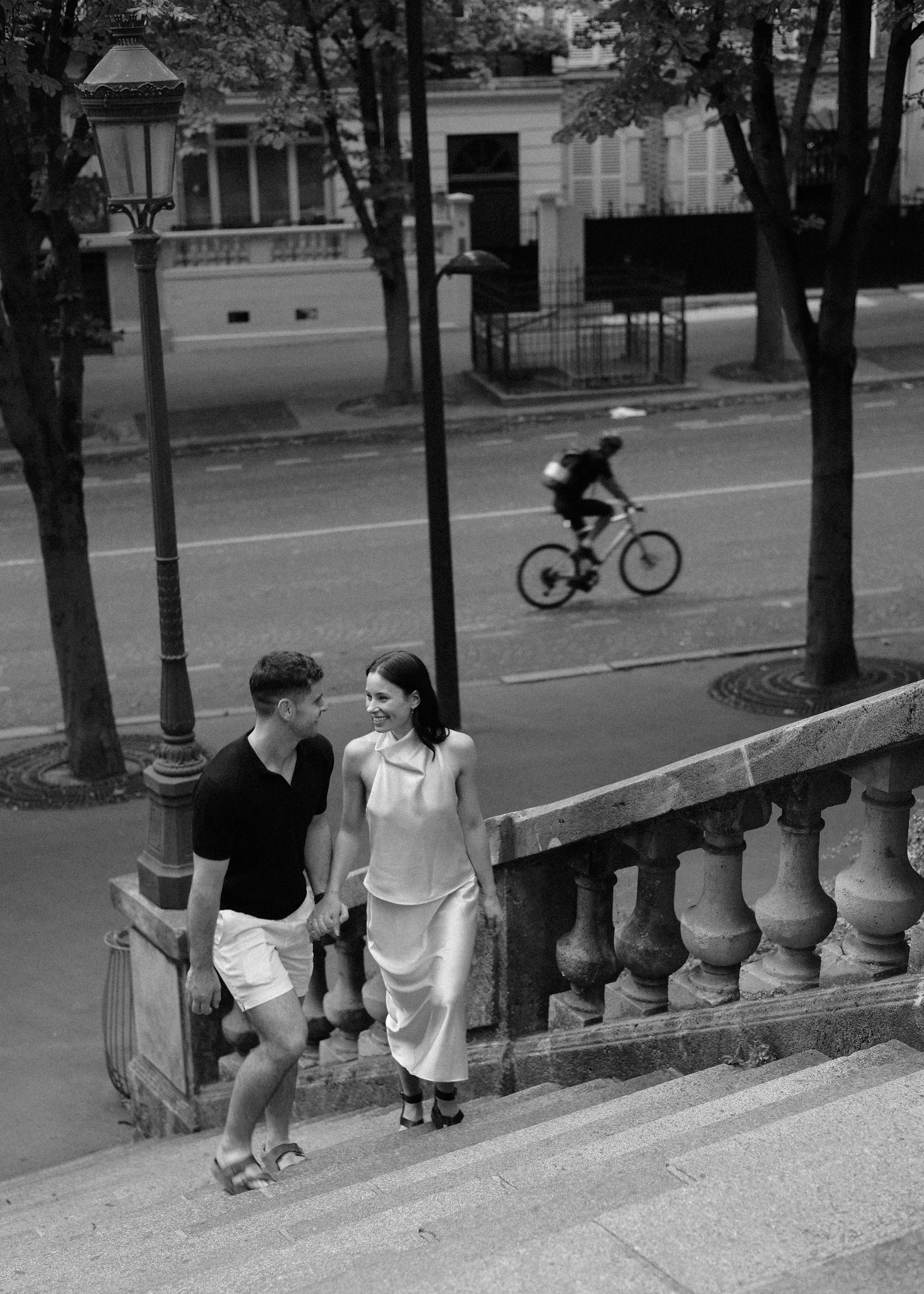 A black and white photo of a smiling couple ascending outdoor stairs, holding hands, with a street scene in the background. A cyclist is riding past on the street behind them.