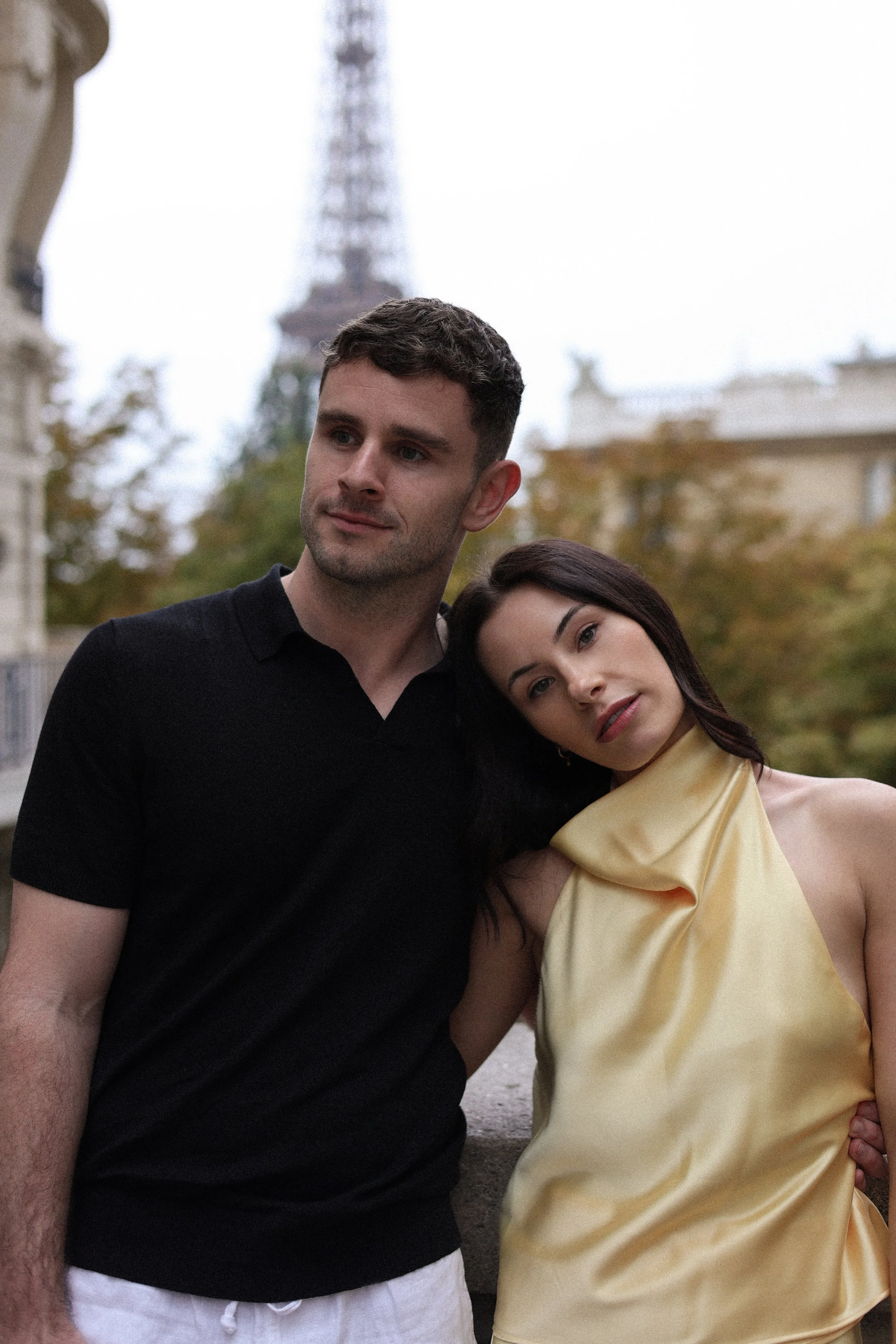 A young man and woman are standing together outdoors, with the Eiffel Tower visible in the background. The man is wearing a black shirt and white pants, and the woman is wearing a yellow satin dress. The woman is resting her head on the man's shoulde