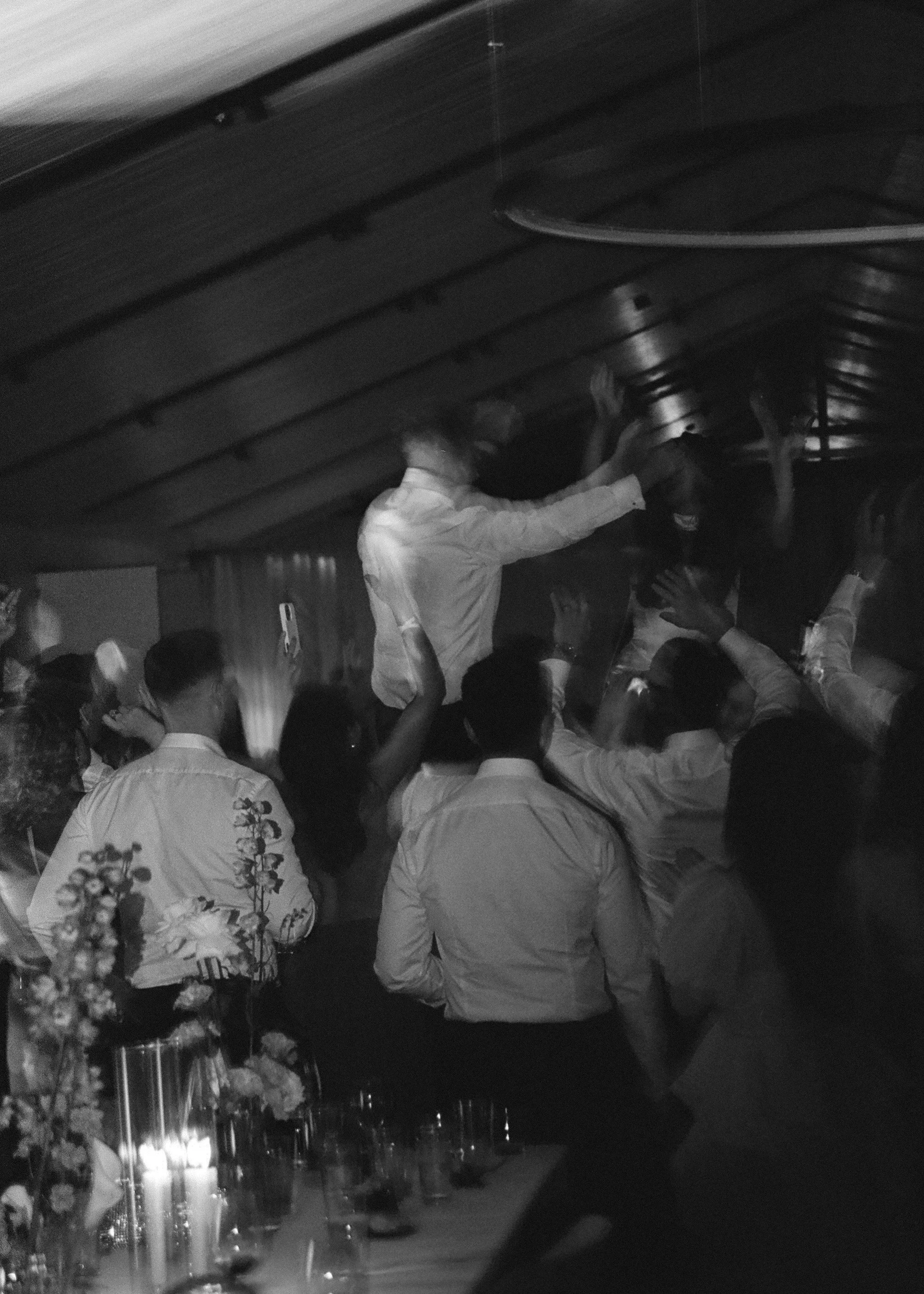 A group of men in formal attire dancing and celebrating at a party, with some raising hands and a man in the center reaching out.