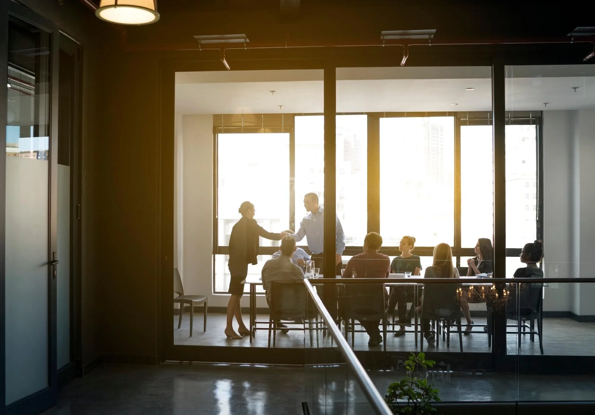Professionals in a sunlit glass-walled conference room, two colleagues shaking hands — reflecting the high-pressure BigLaw environment that drives attorney burnout, workaholism, and the need for lawyer mental health support