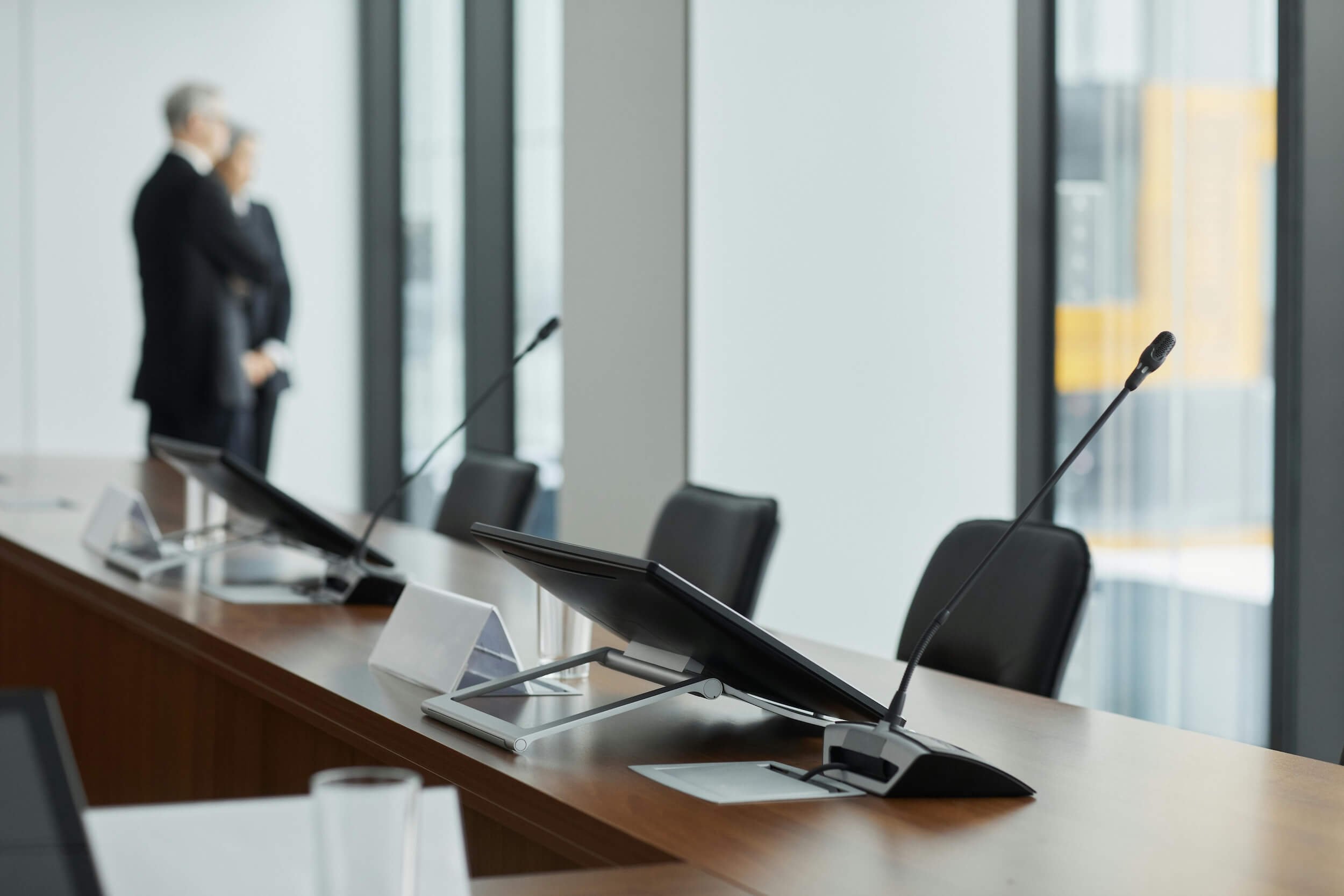 Empty boardroom conference table with microphones and monitors, two professionals in conversation by floor-to-ceiling windows — representing the high-pressure legal environment that lawyer therapy addresses