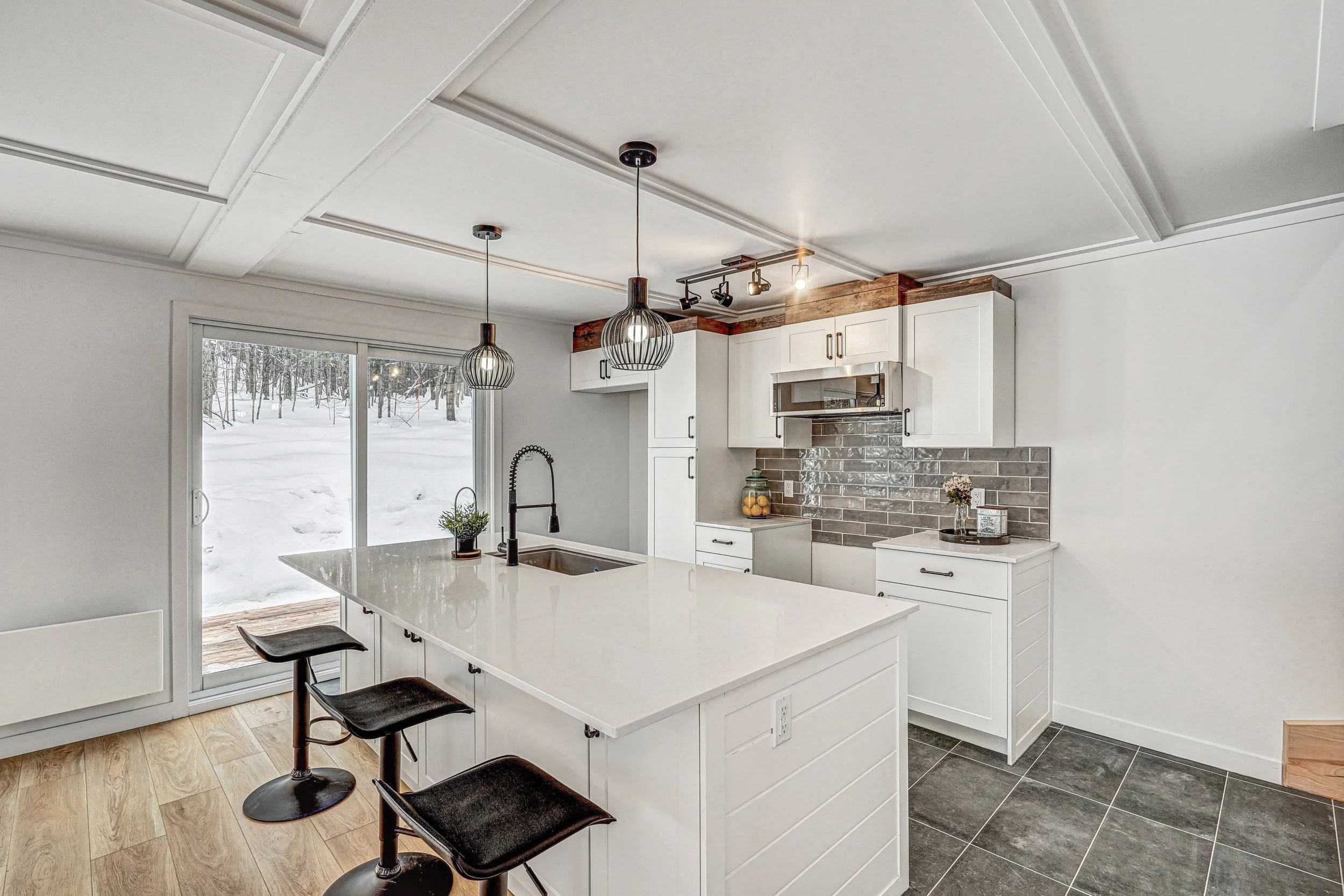 Cuisine moderne avec îlot central, comptoirs en quartz blanc, murs en brique grise, éclairage suspendu et vue sur un extérieur enneigé par une grande fenêtre.
