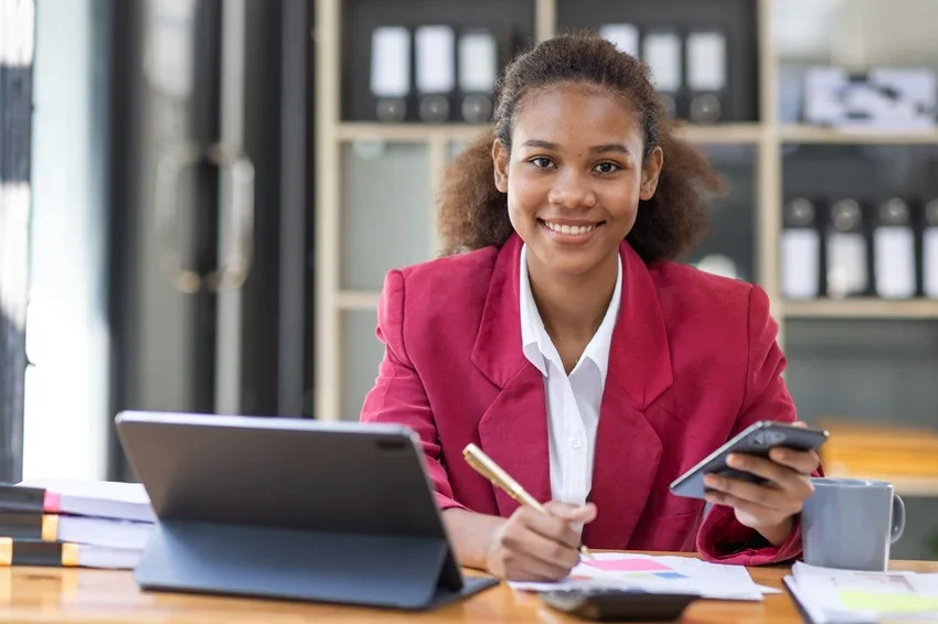 Cedeno & Associates | Nonprofit Tax & Accounting Services – Smiling woman in pink blazer sitting at a desk with a tablet and smartphone, in an office setting.