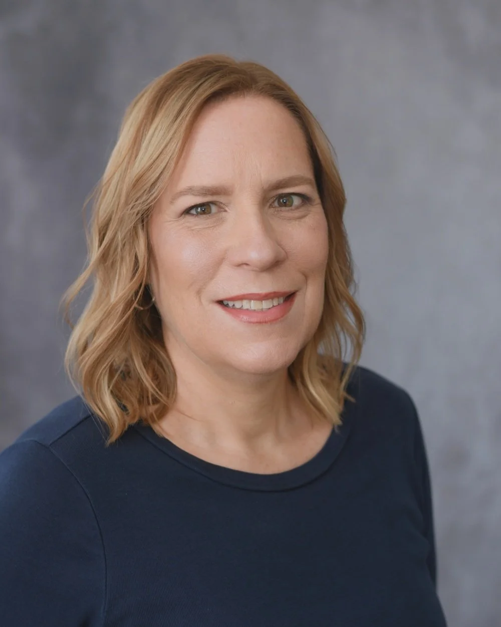 A woman with shoulder-length wavy blond hair, fair skin, and brown eyes, smiling at the camera, wearing a navy blue top, against a gray background.