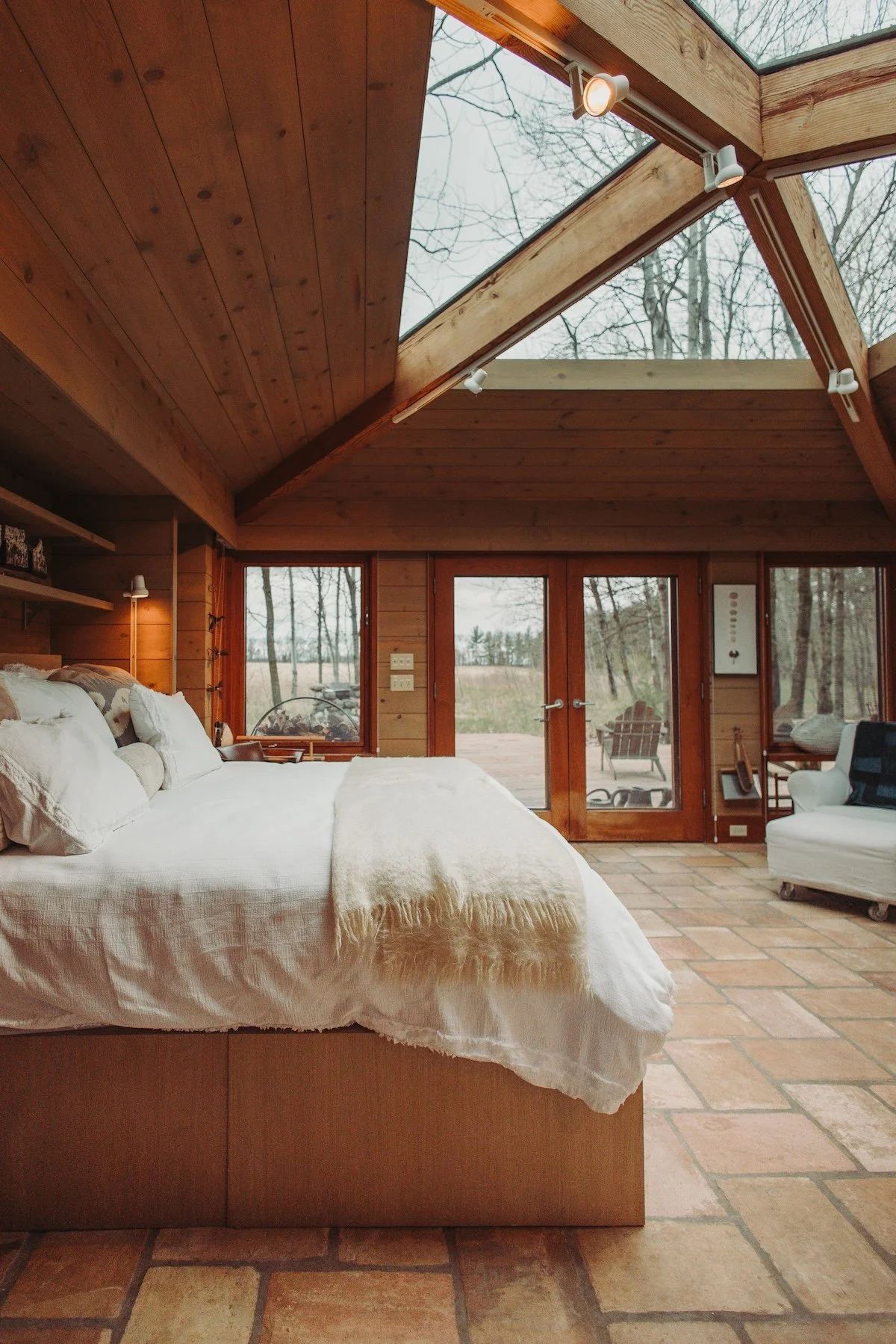 Interior of a cozy bedroom with wooden walls and ceiling, a large bed with white bedding, and glass doors leading to an outdoor deck with chairs, set in a wooded area.