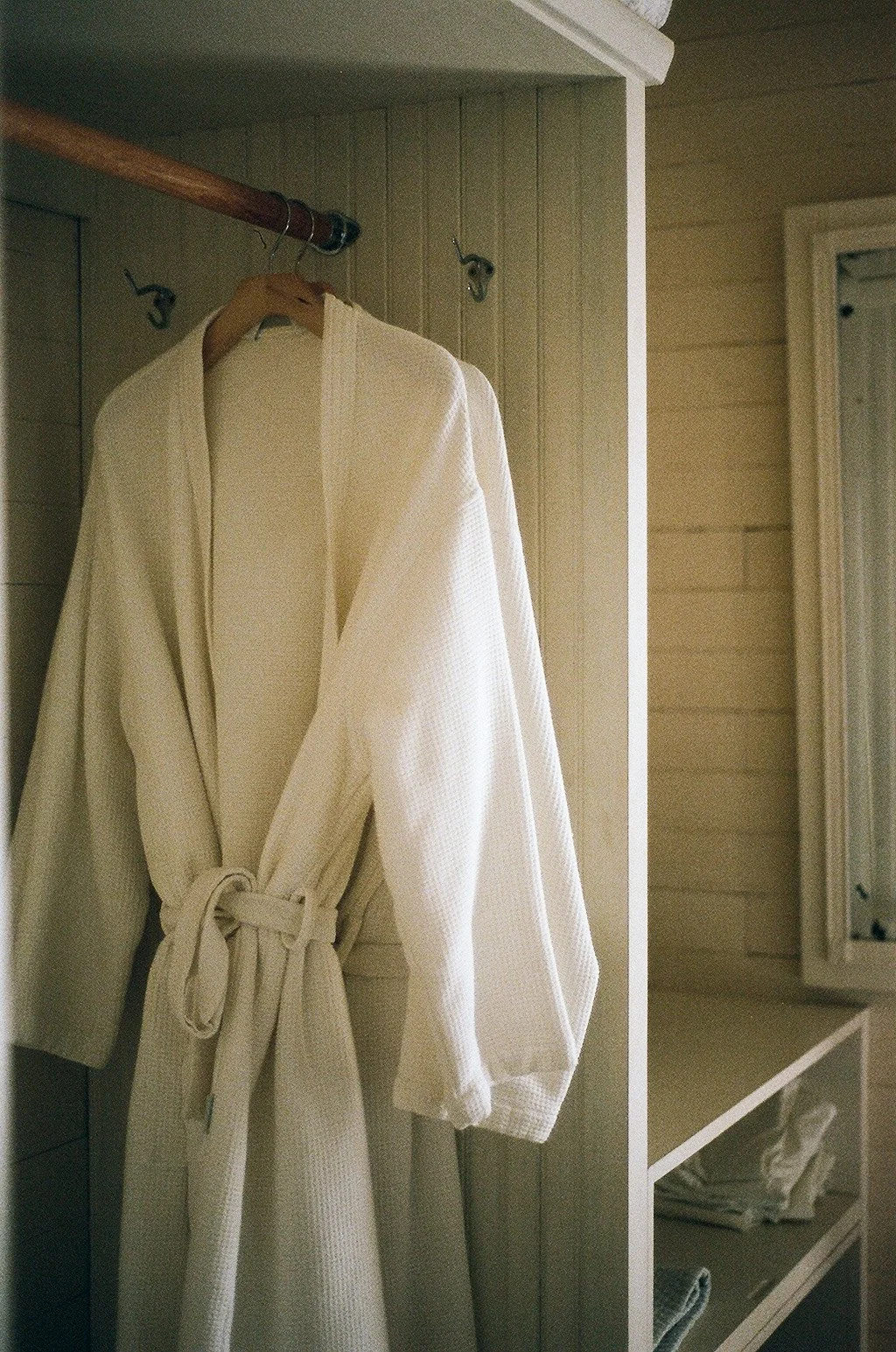 A white bathrobe hanging on a hook inside a closet with open shelves and a window