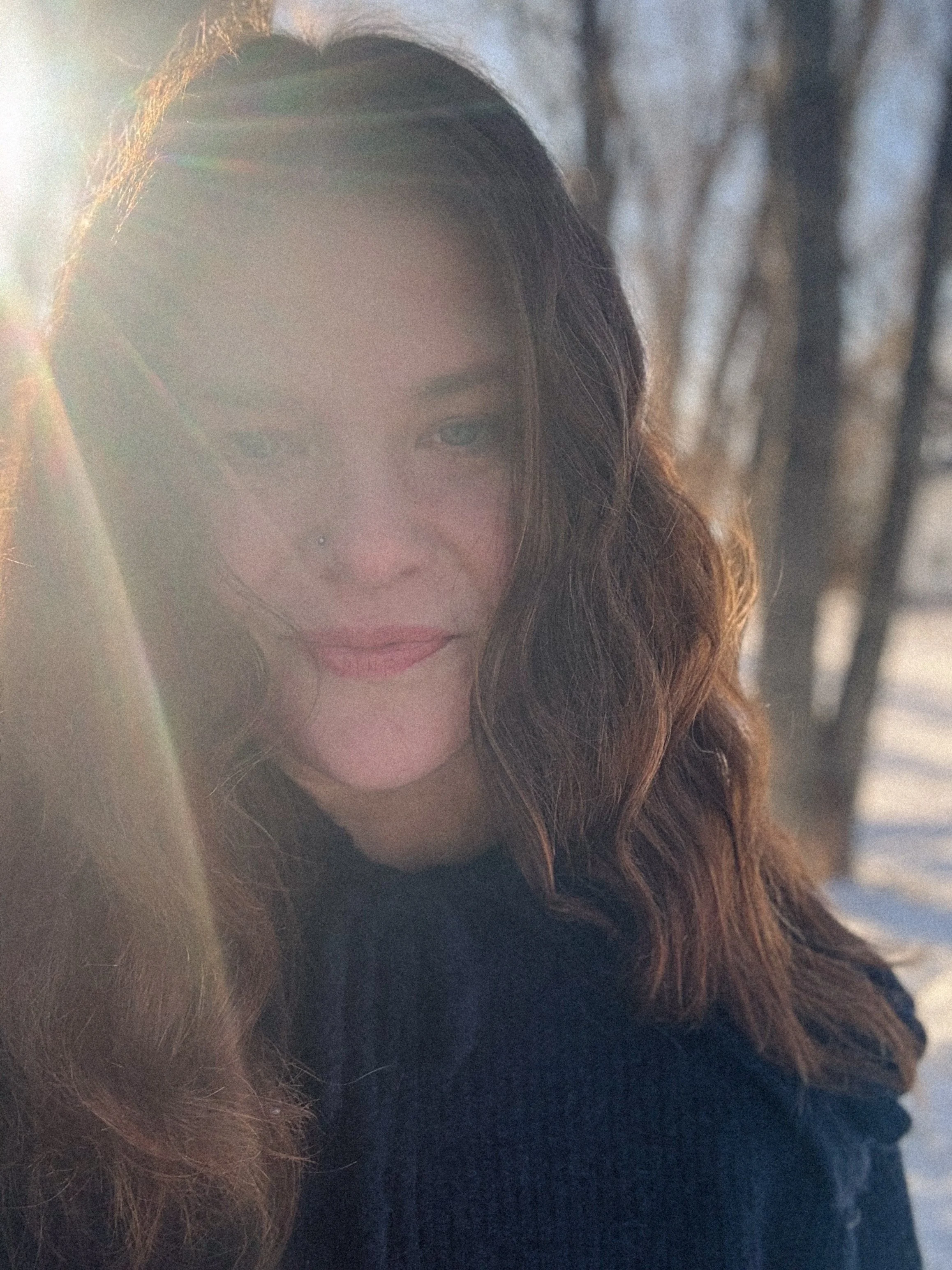A woman with wavy brown hair and a nose piercing smiling outdoors with sunlight shining on her face, winter trees in the background.
