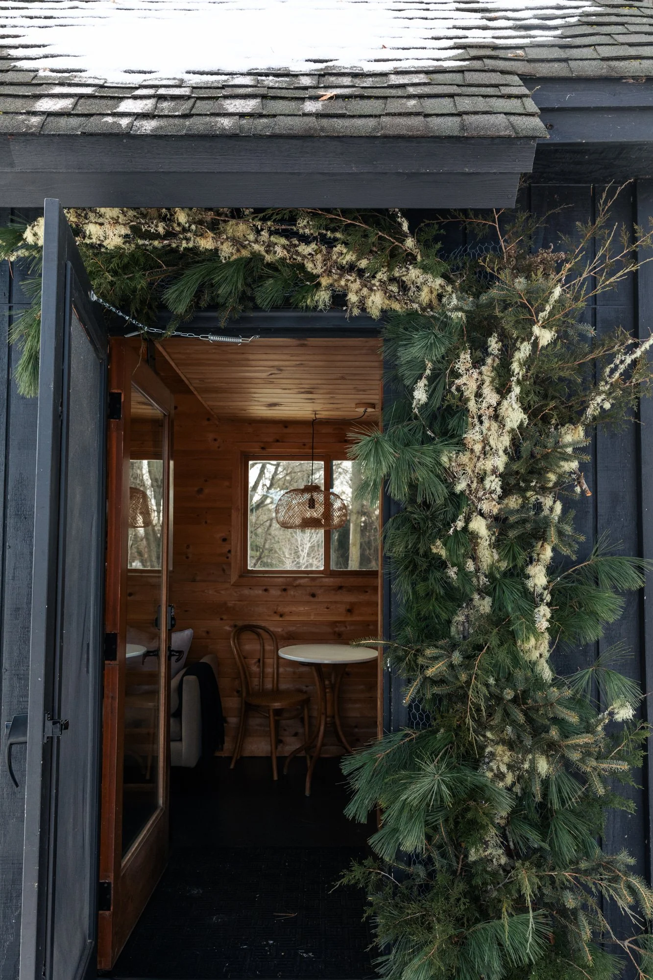 Black wooden exterior with snow on the roof, decorated with green pine branches and white flowers, and string lights hanging above a glass door.