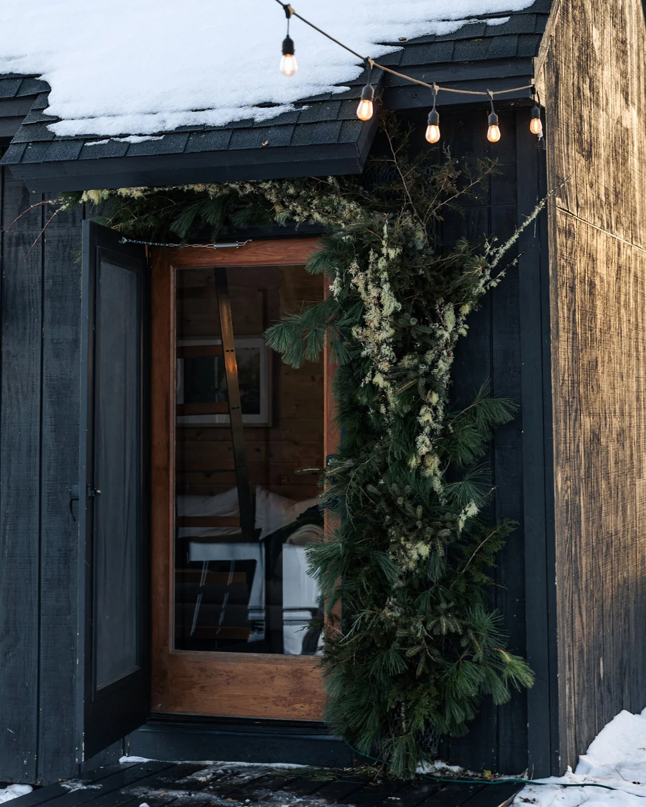 Black wooden exterior with snow on the roof, decorated with green pine branches and white flowers, and string lights hanging above a glass door.