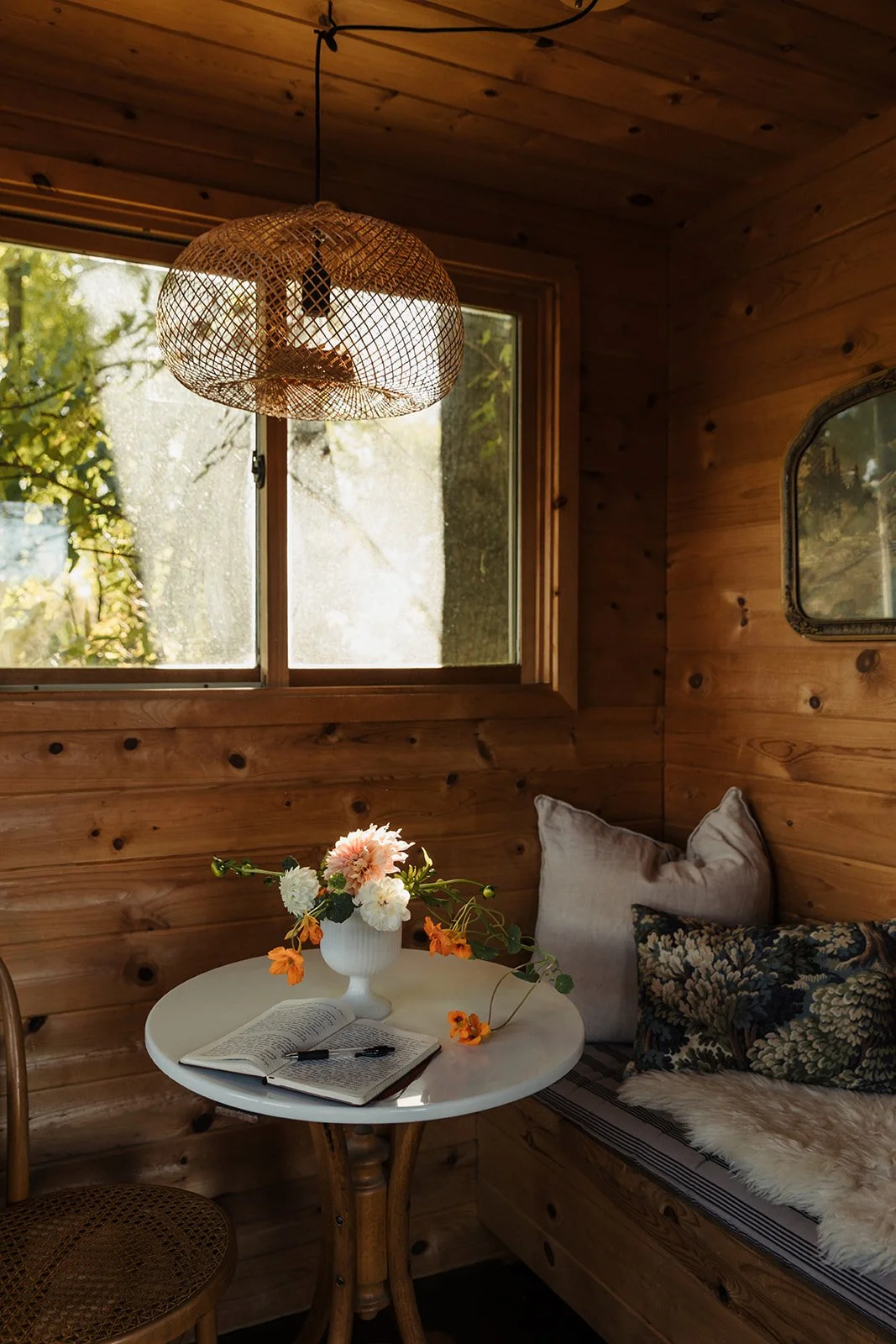 A cozy wooden interior with a window showing greenery outside, a round table with a vase of flowers, an open book, and cushions on a seating area.
