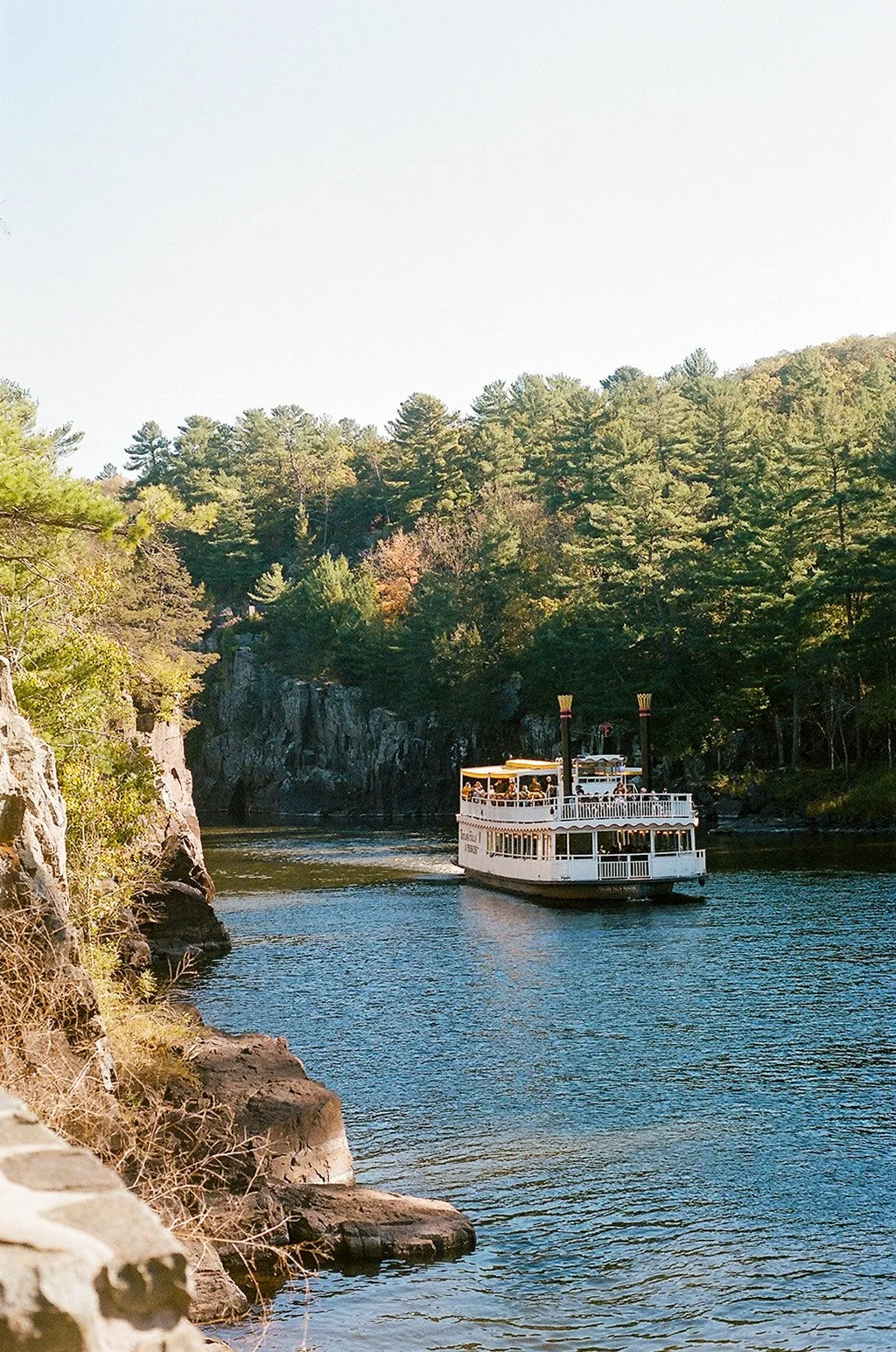 A boat with multiple decks sailing on a river surrounded by lush, green trees on a clear day.