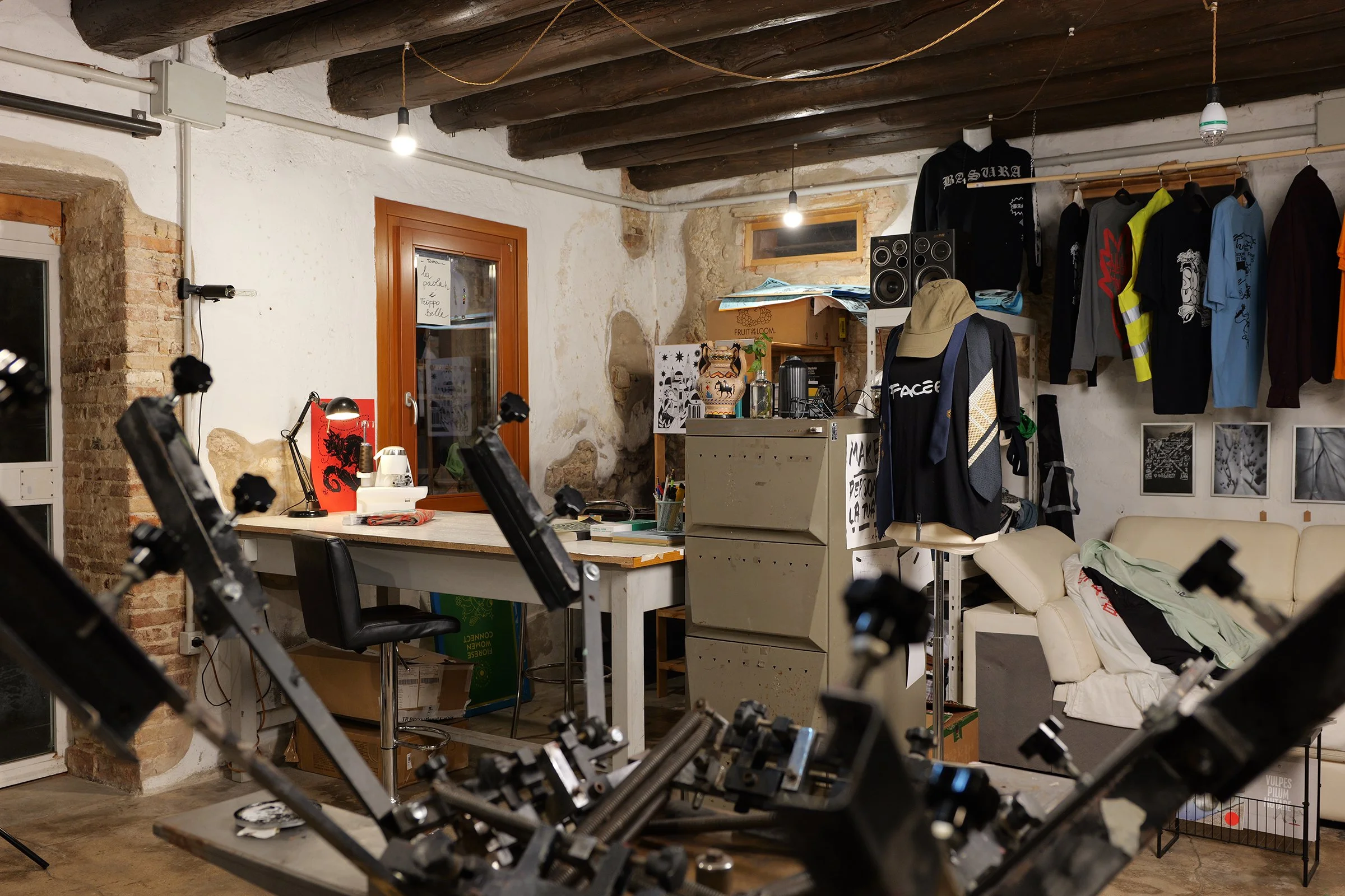 An indoor room with a cluttered workspace and various objects. In the foreground, there are robotic arms or machinery parts. The background has a white wall with exposed brick and wooden ceiling beams. There are clothes hanging on a rack, a beige sofa with a blanket, books, and miscellaneous items, and a small window.