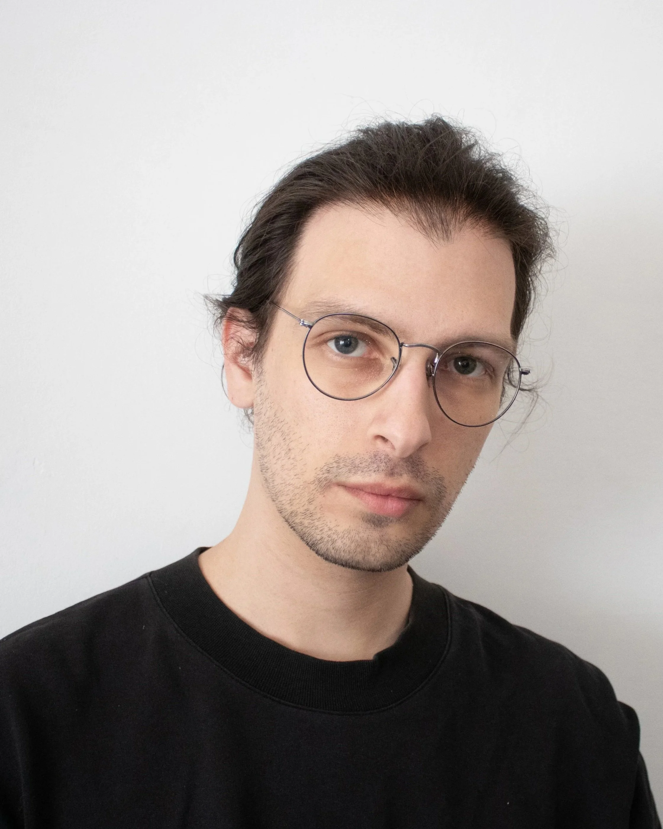 A young man with dark hair, wearing glasses with thin round frames, a black crewneck shirt, and a light stubble, stands against a plain white wall.
