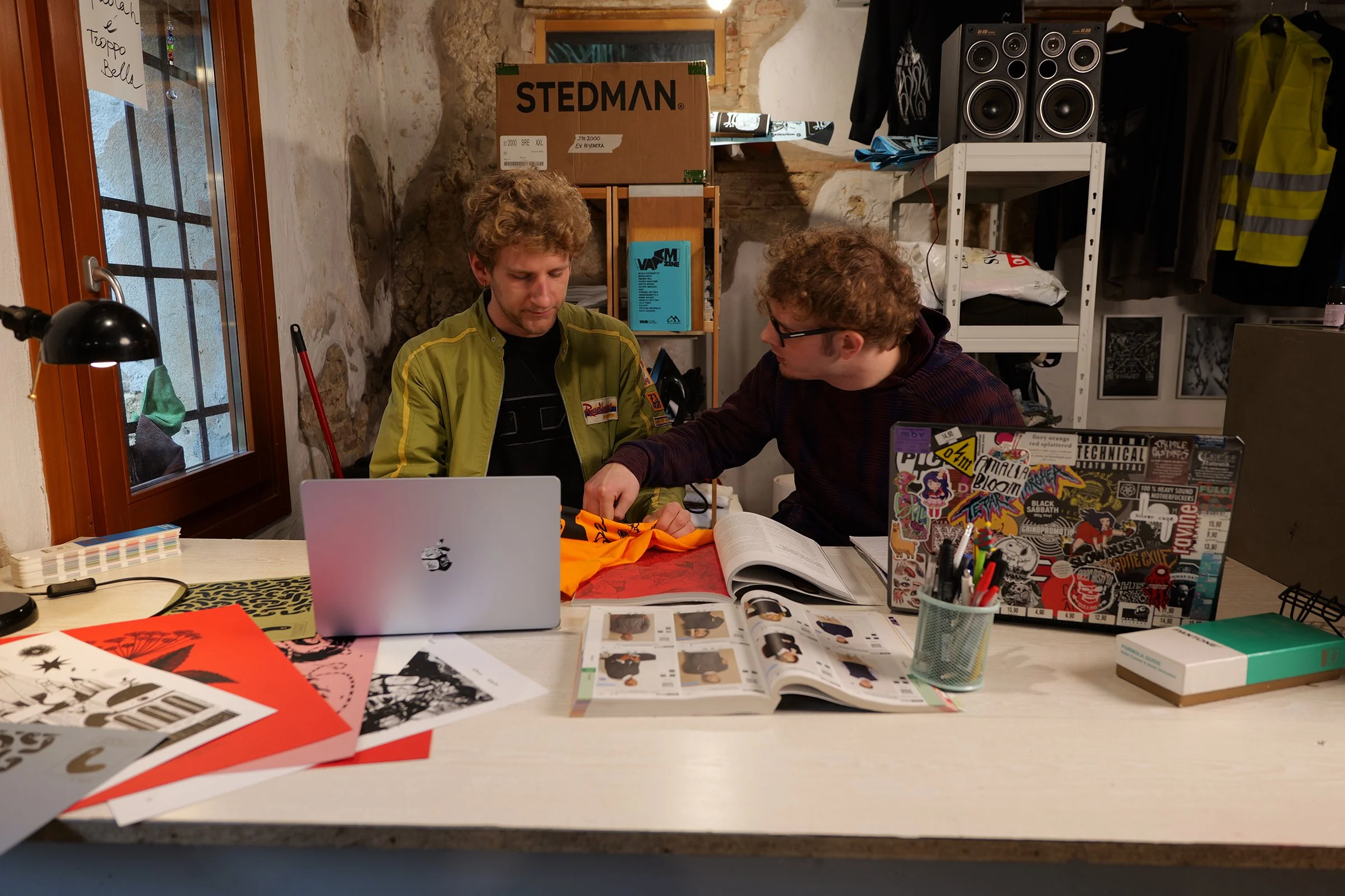 Two young men sitting at a cluttered desk in a room with stone walls, working together on a project. One is pointing at a book, the other is looking at the book and a laptop covered with stickers.