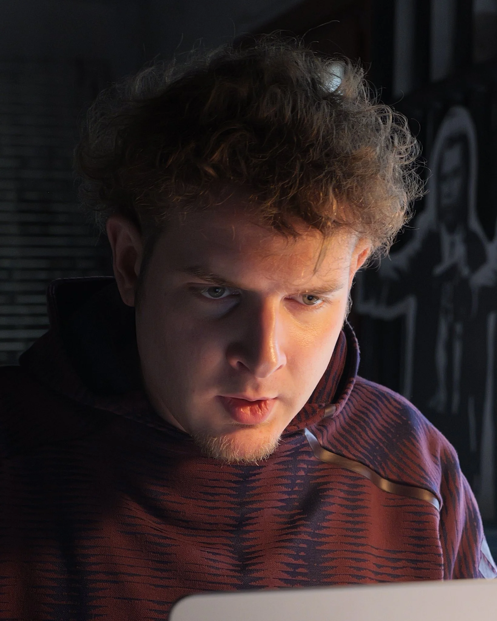 A young man with curly hair looking at a screen with a serious expression in a dimly lit room.