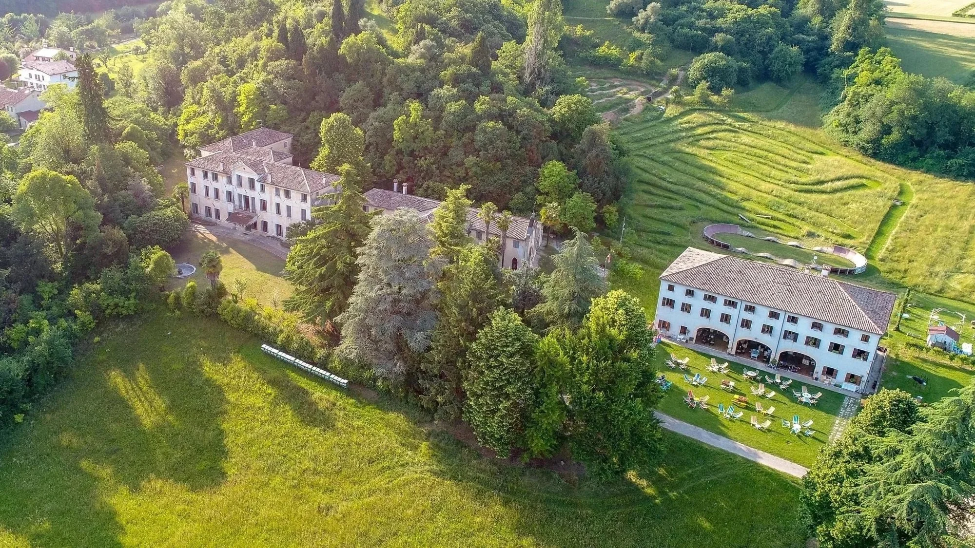 Aerial view of a large estate with multiple buildings surrounded by lush green trees, manicured lawns, and a hilly landscape, including a small circular area with outdoor seating.