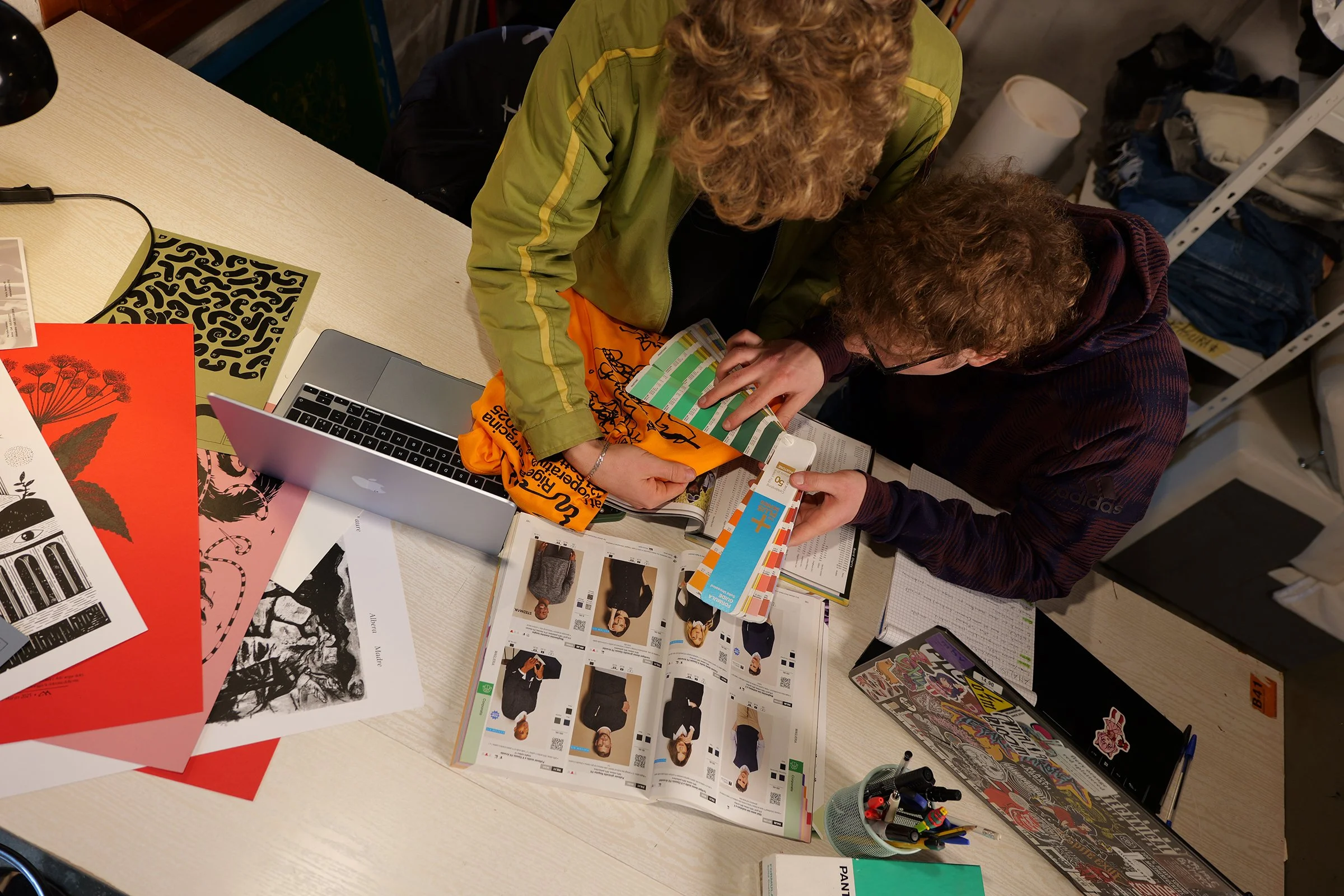 Two people are studying or working at a desk covered with colorful papers, a laptop, a magazine, and various stationery items.