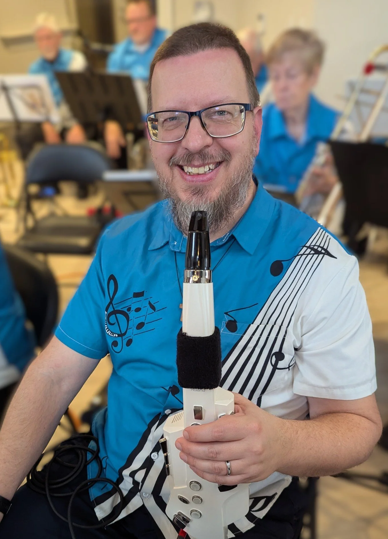 A man with glasses and a beard playing a recorder, smiling and sitting in a room with other people.