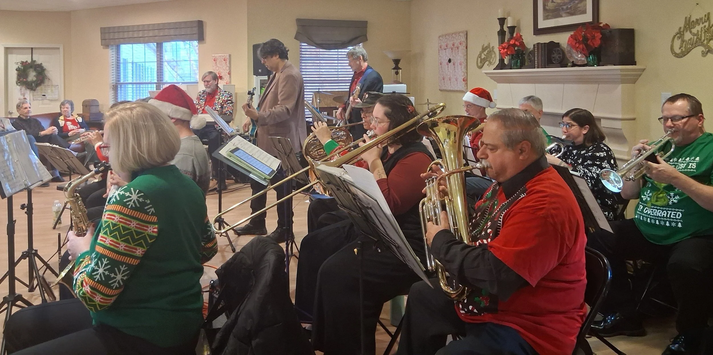 A group of musicians performing in a Christmas-themed concert. Some are wearing holiday sweaters and Santa hats, and the scene is decorated with holiday wreaths and poinsettias.