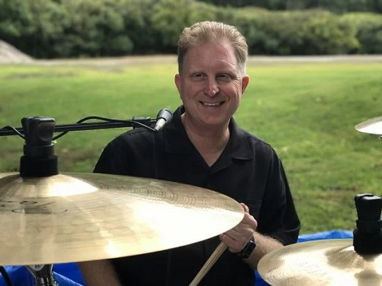 A smiling man with light hair wearing a black shirt, playing drums outdoors on a grassy field with trees in the background.