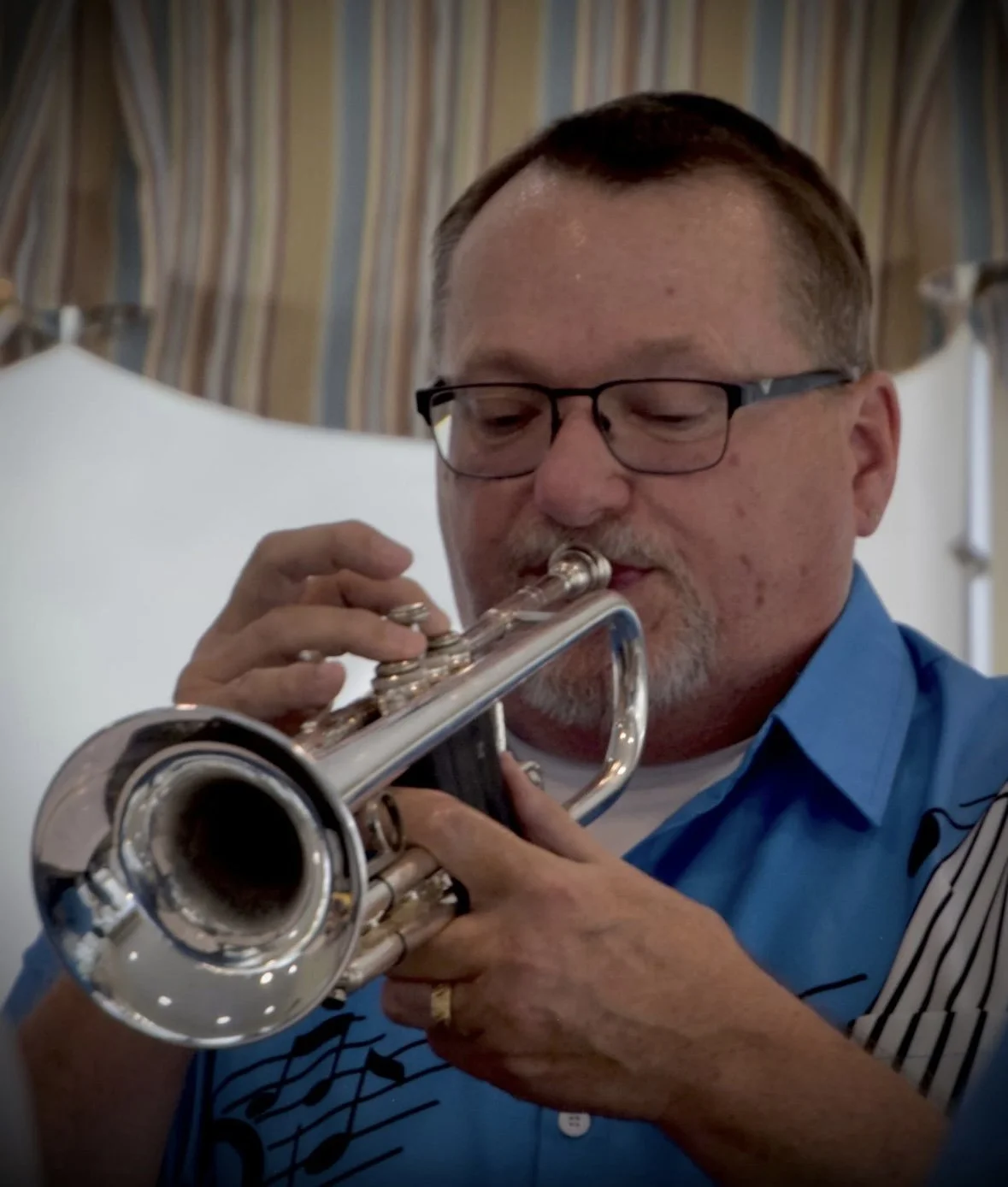 Man playing trumpet, wearing glasses and a blue shirt, in indoor setting with curtains in background.
