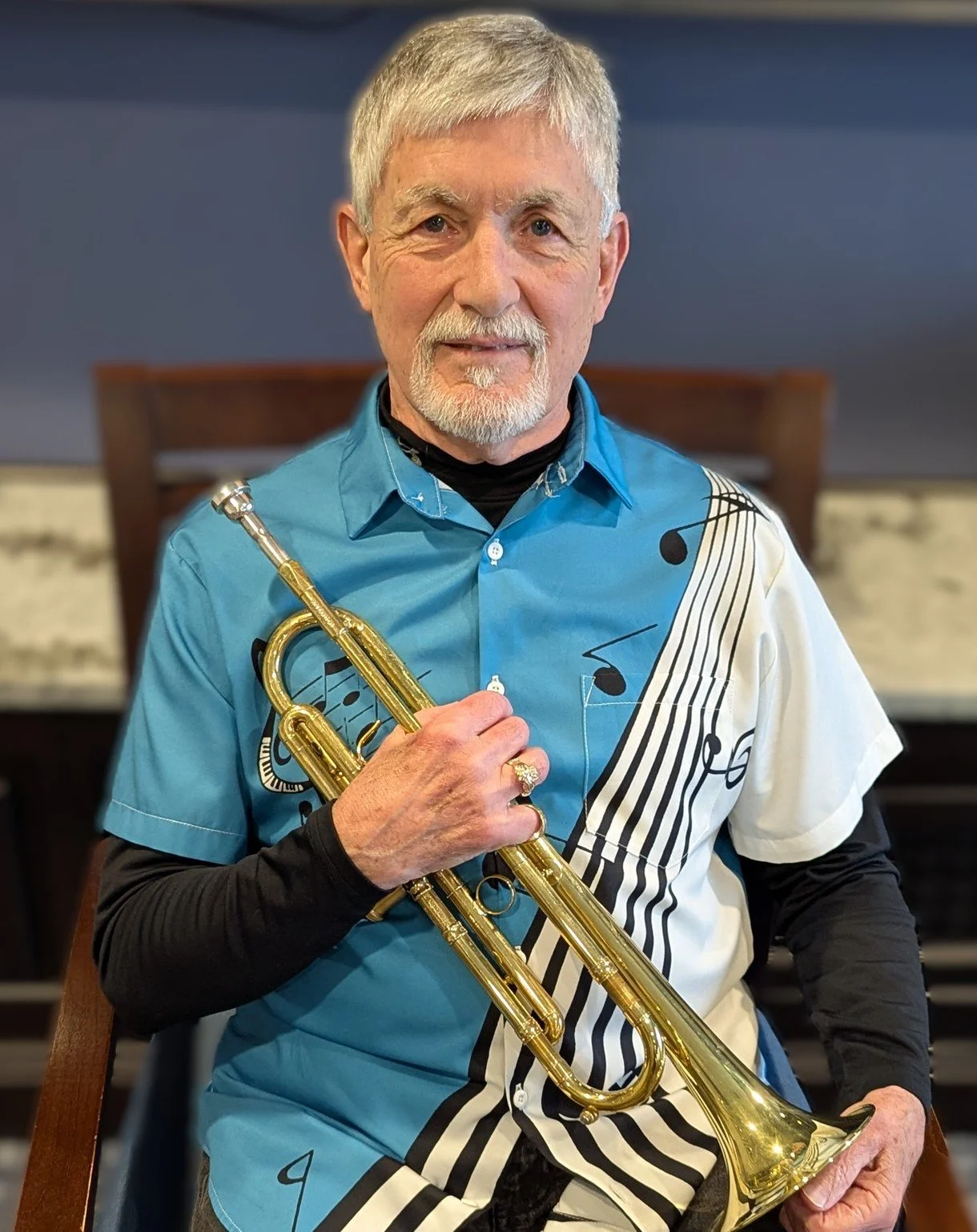 Older man wearing sunglasses and a white T-shirt with an American flag graphic, holding a camera or telescope, standing outdoors with green foliage in the background.