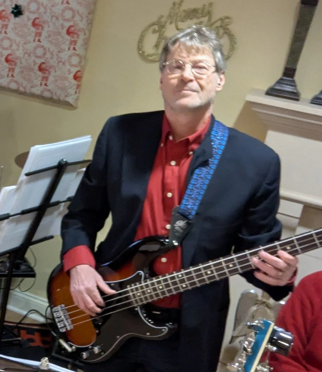 A man wearing glasses, a red shirt, and a black jacket playing an electric bass guitar at an indoor event.