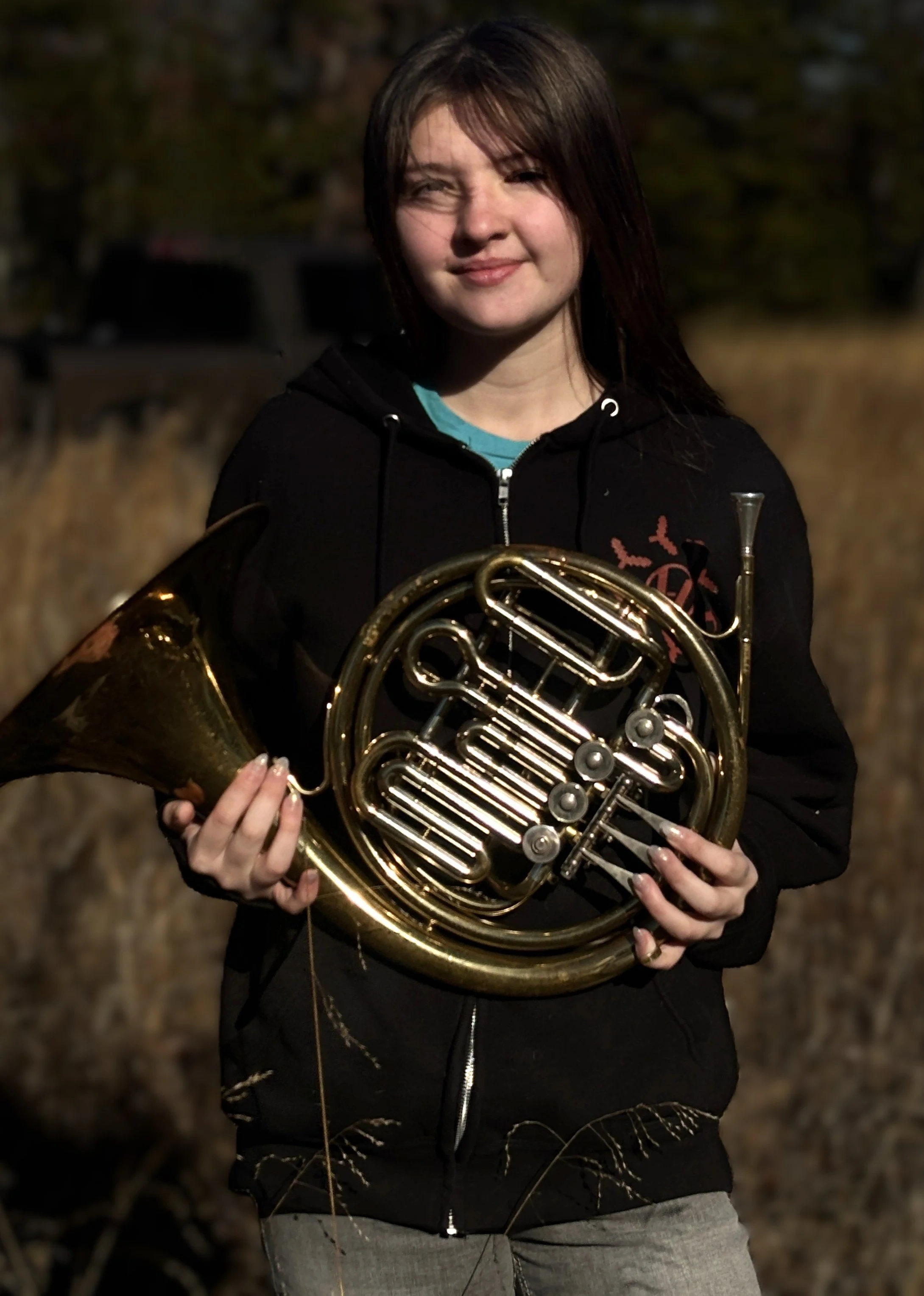 A young woman with brown hair holding a brass French horn outdoors at dusk, wearing a black hoodie and smiling.