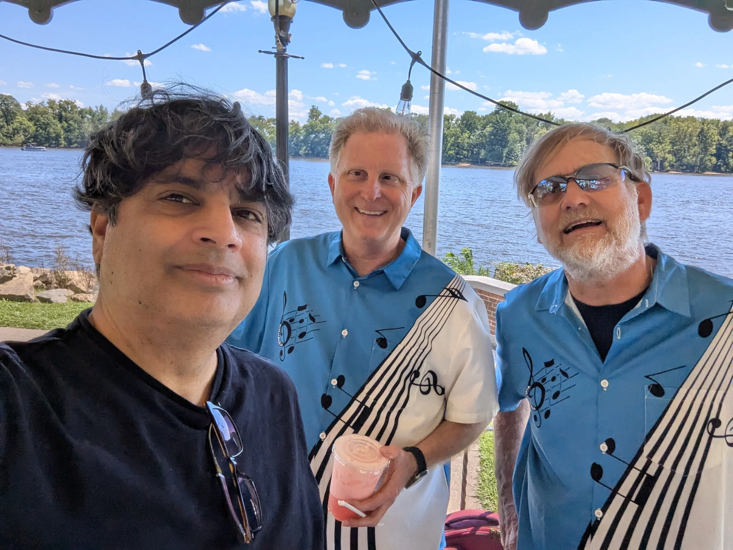 Three men taking a selfie outdoors by a body of water with trees in the background. The man on the left has dark curly hair and is wearing a black shirt. The man in the middle has short light hair and is holding a pink smoothie. The man on the right 