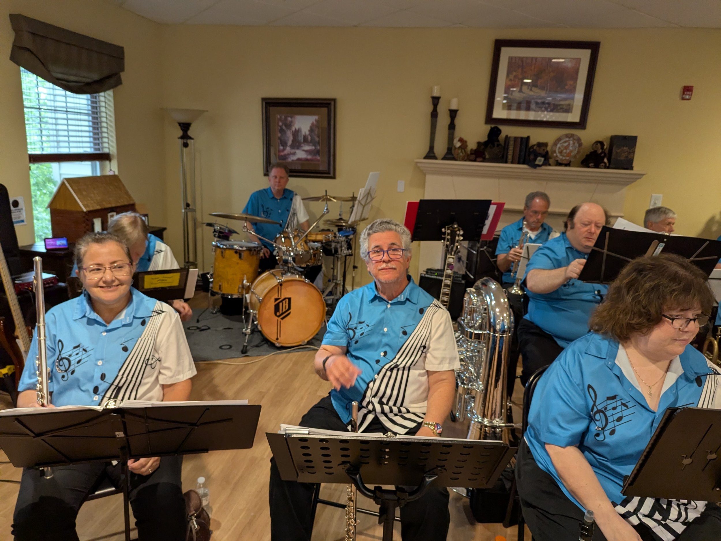 A group of seniors in matching blue and white shirts with musical notes, holding instruments, in a living room setting with a fireplace, framed pictures, and musical equipment.