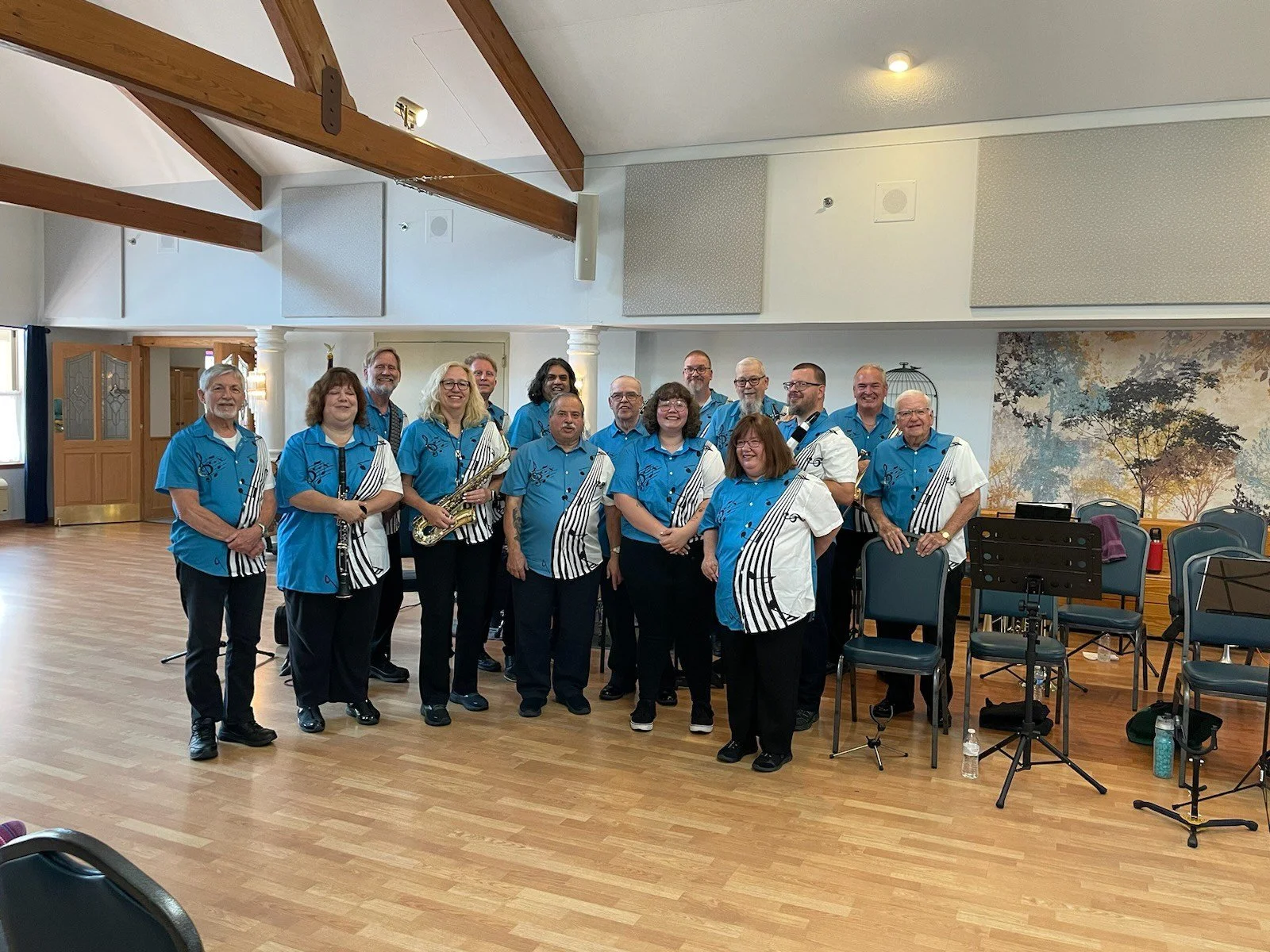 Group of 16 people, mostly middle-aged or older, standing in a spacious room with wooden beams and a large mural of trees on the wall. They are dressed in matching blue and white shirts, some holding musical instruments like saxophones. There are cha