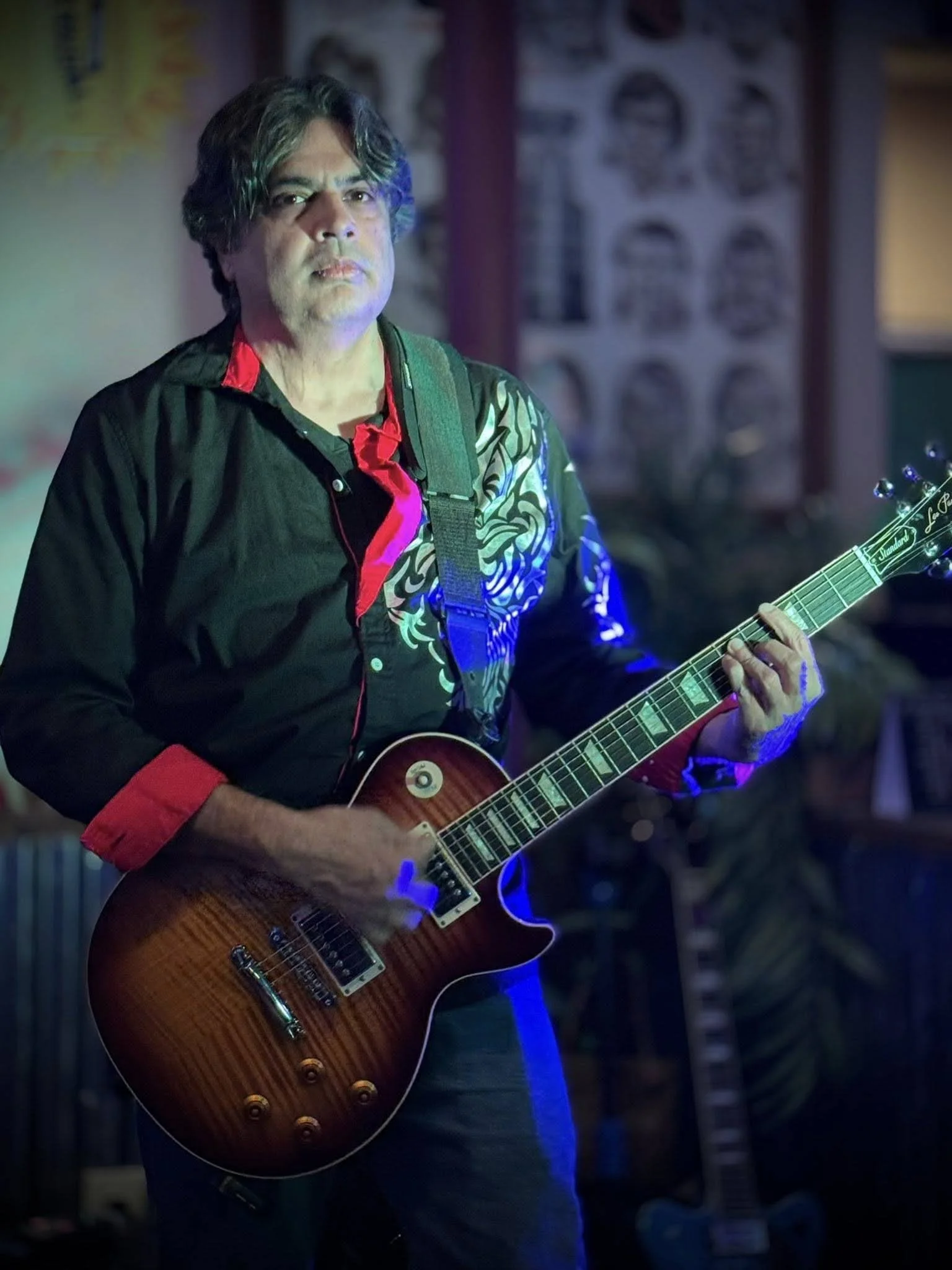 A man with long dark hair, wearing a black t-shirt, sitting with an electric guitar in a guitar store or studio, with several other guitars hanging on the wall behind him.