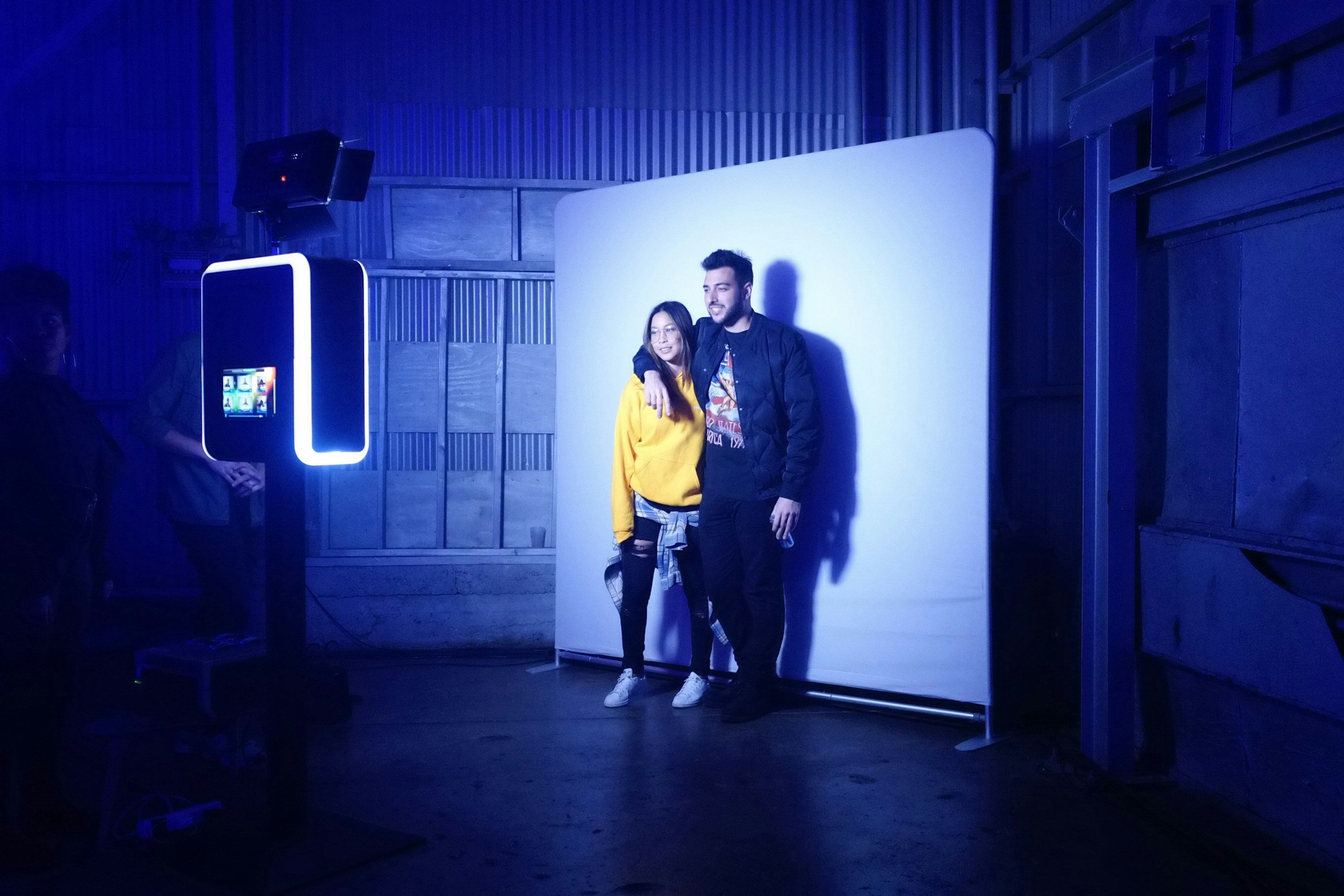 Two people posing in front of a white backdrop, illuminated with blue lighting, in a dark indoor setting with a photo booth on the left.