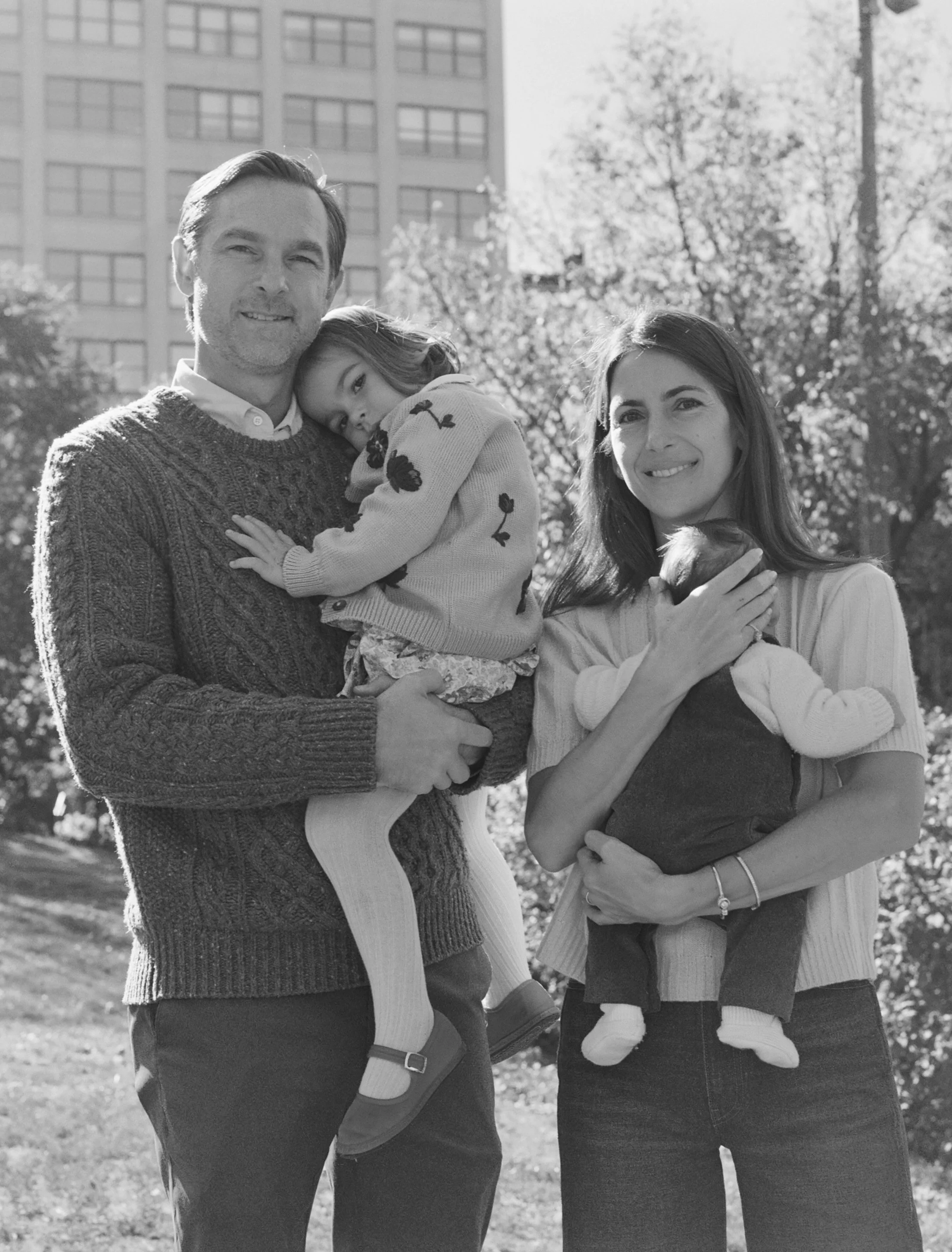A black and white photo of a family of four outdoors, with a man, woman, and two young girls, holding the girls in their arms and smiling.