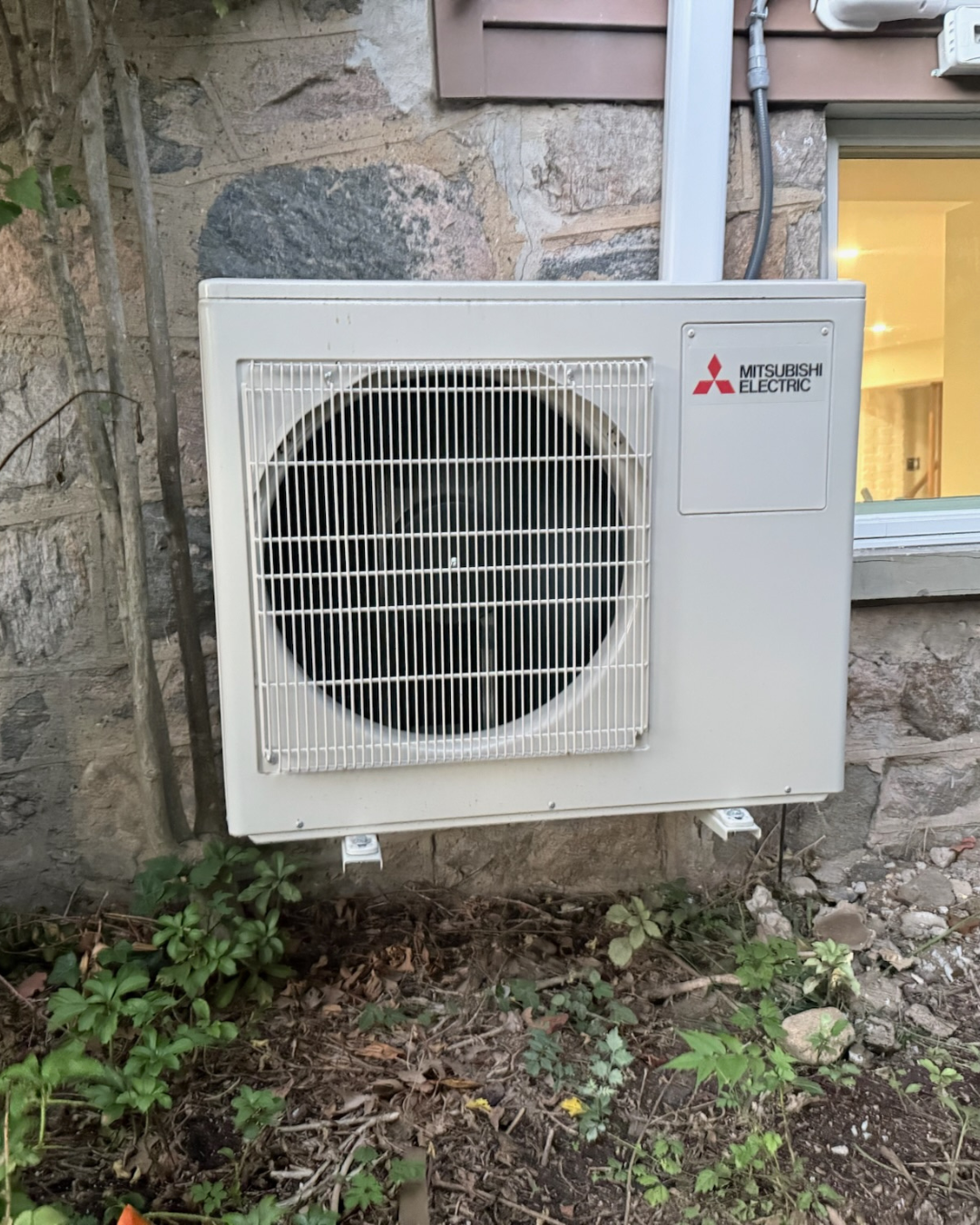 Mitsubishi Electric outdoor air conditioning unit mounted on stone wall outside of house, with window nearby and some small plants growing below.
