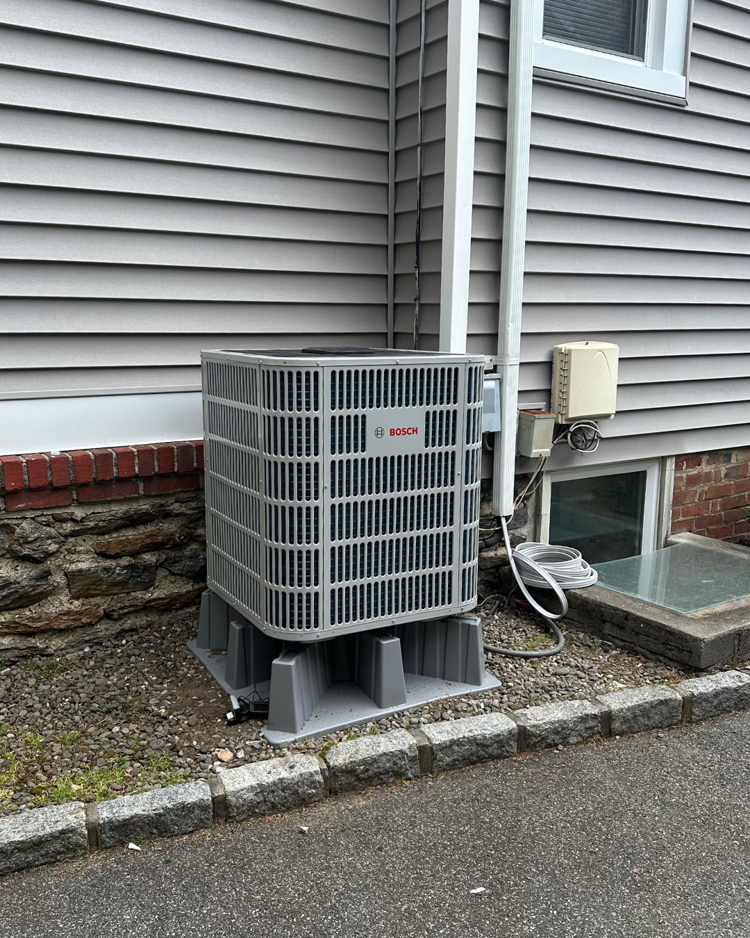 Outdoor view of a HVAC unit on concrete pad next to house with siding, window, and utility connections.