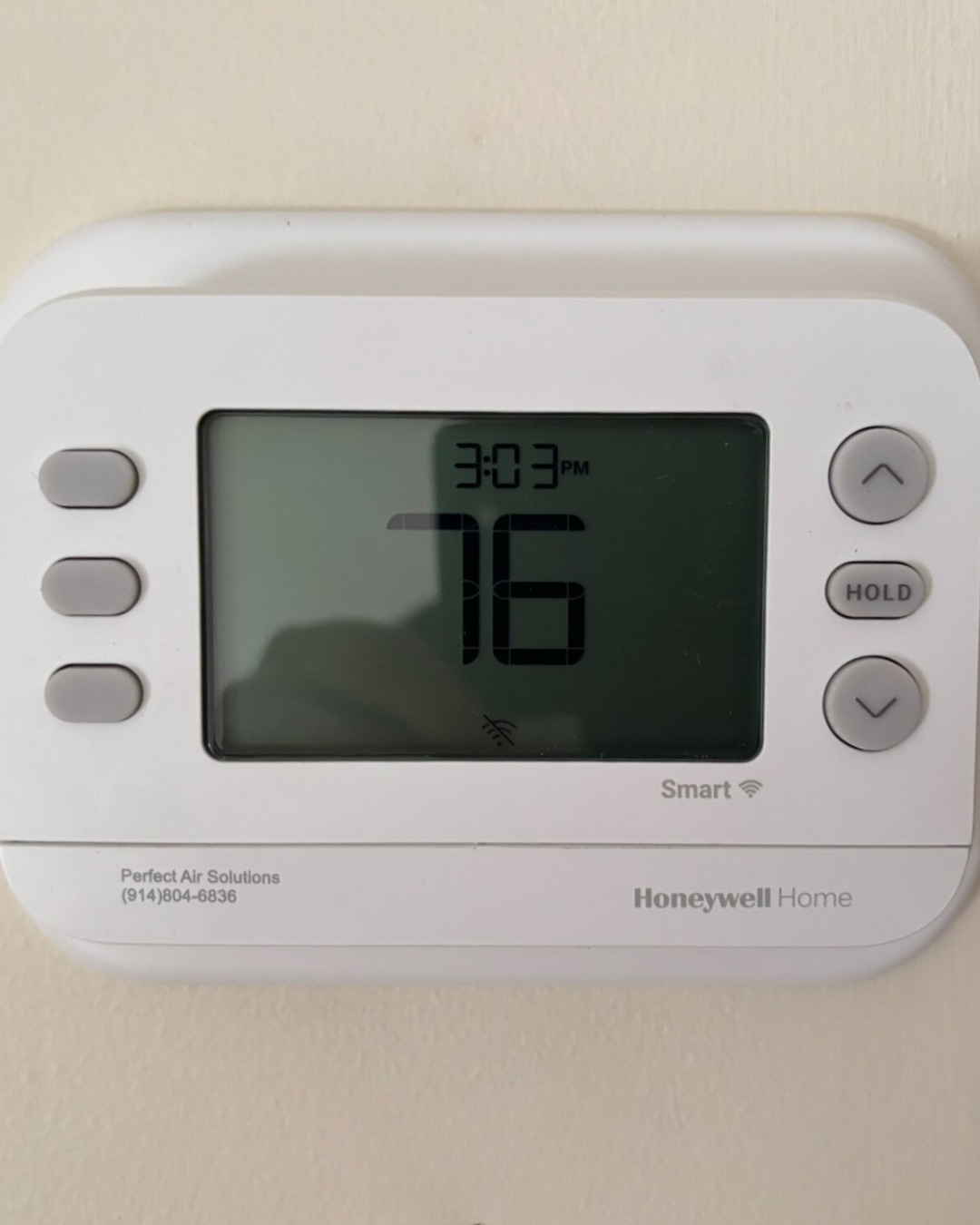 Digital thermostat showing 76 degrees Fahrenheit, time 3:03 PM, mounted on a wall with beige paint.
