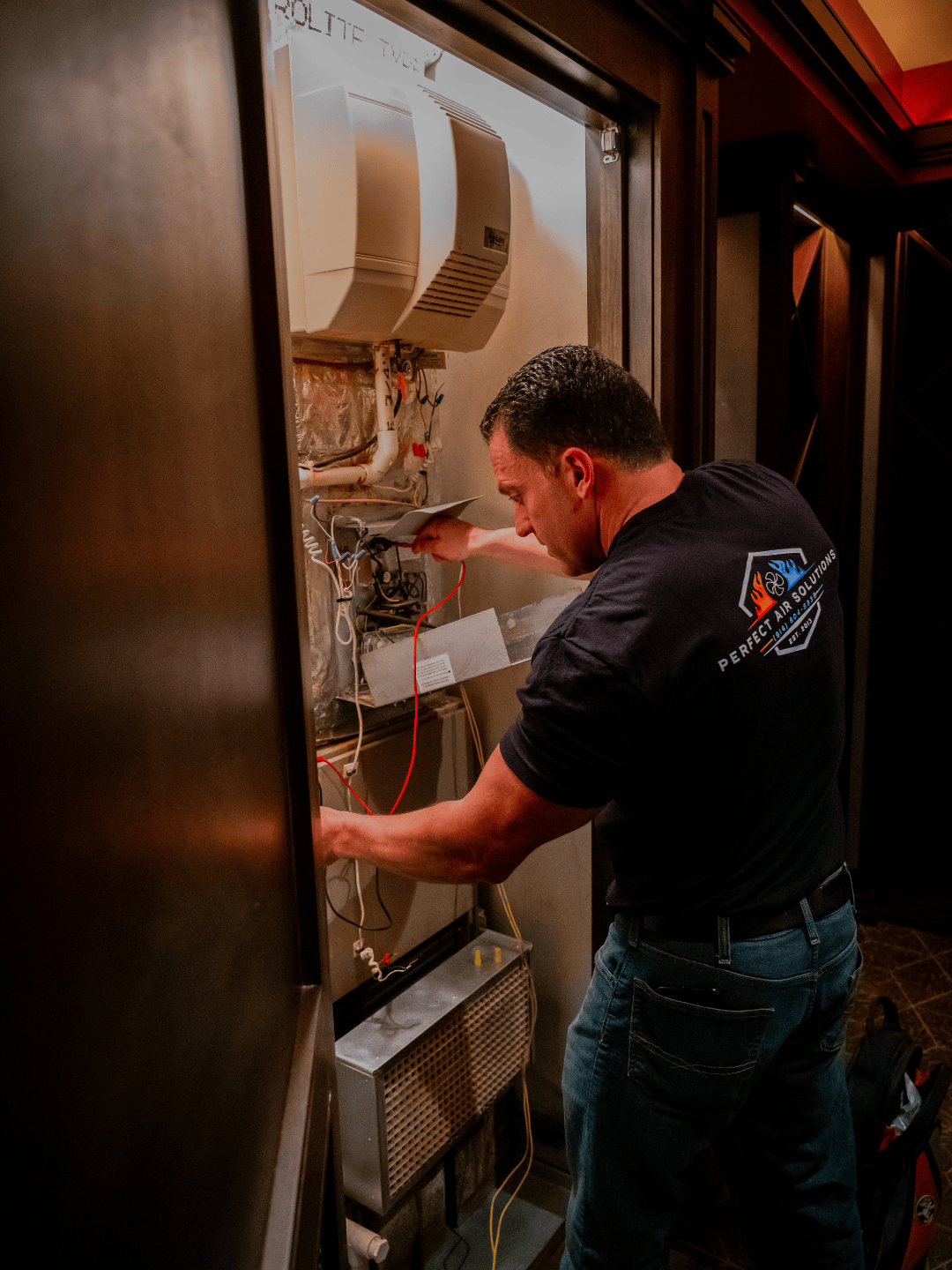 A technician working on repairing or maintaining an electrical or HVAC system inside a cabinet, wearing a black T-shirt with a logo for 'Perfect Air Solutions'